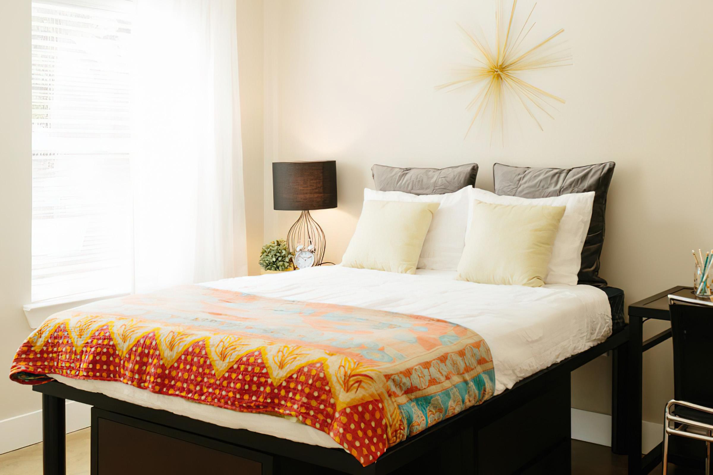 A cozy bedroom featuring a bed with a colorful patterned throw, two light-colored pillows, and stylish gray headboards. There's a modern lamp on a bedside table, a small potted plant, and a sunburst wall clock accentuating the warm, inviting atmosphere. Natural light filters through sheer curtains.