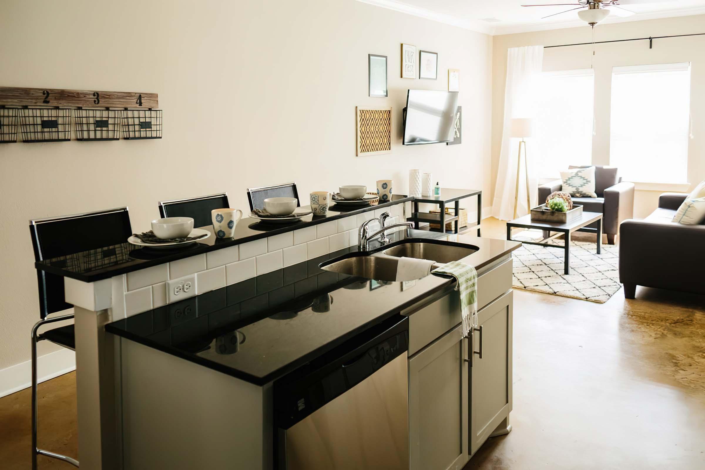 Modern kitchen with a black granite countertop and stainless steel appliances. The kitchen island features two bar stools and is set with dishes. Nearby, a cozy living room is visible, featuring a gray sofa, decorative pillows, and a standing lamp. Natural light streams through large windows with sheer curtains.