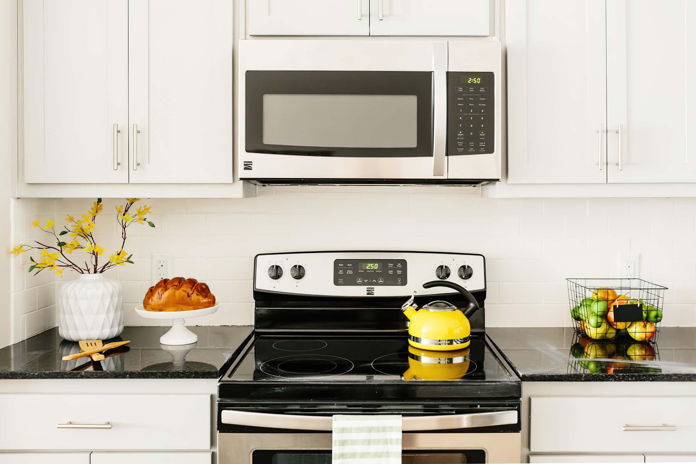 A modern kitchen featuring a sleek black stove with a stainless steel microwave above. A yellow kettle sits on the stove, with a green and white striped towel hanging. To the left, a vase with yellow flowers and a cake on a stand are visible, while a wire basket filled with apples is on the right.