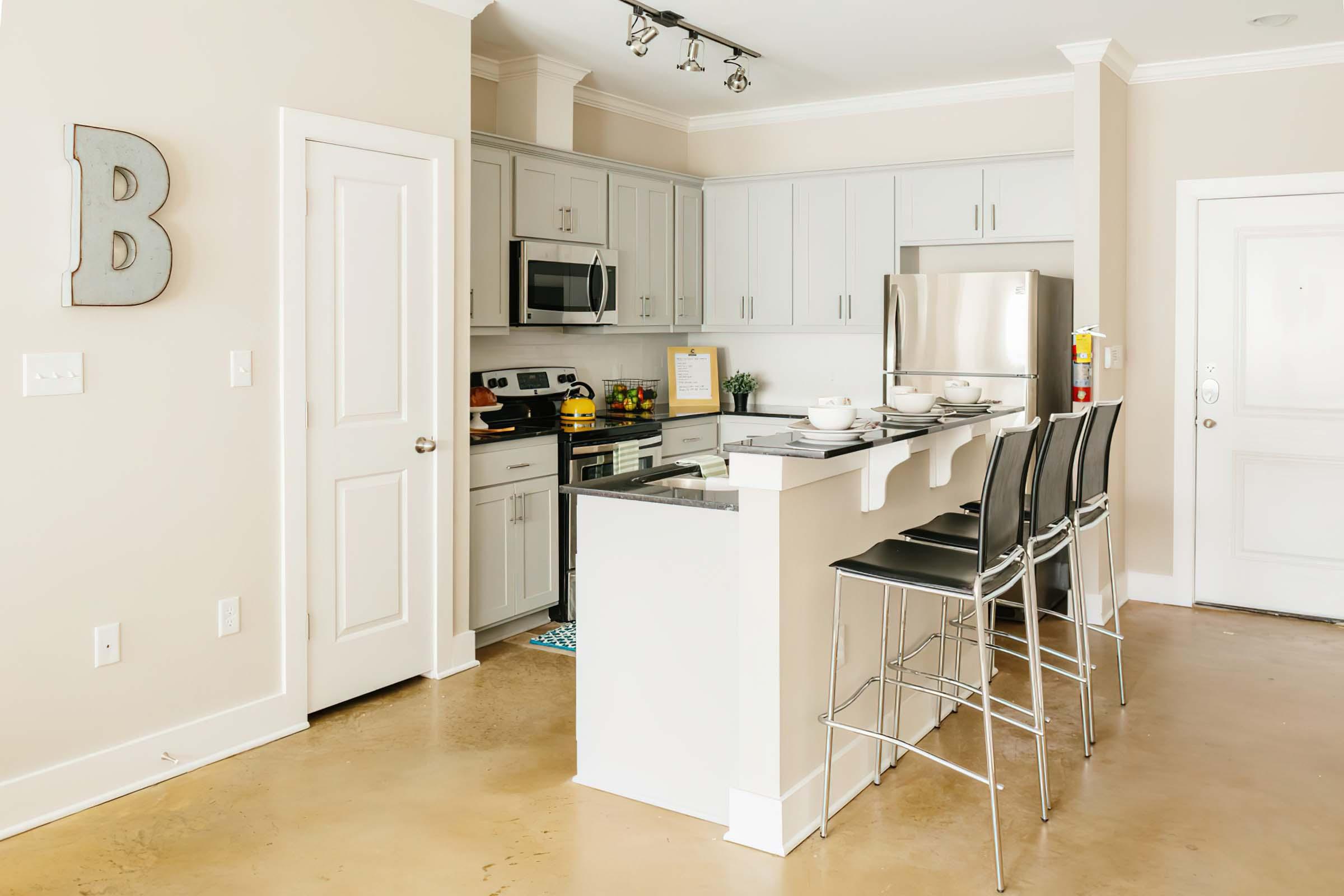 Modern kitchen featuring light gray cabinets, granite countertops, stainless steel appliances, and bar stools. The space is well-lit with recessed lighting and has a neutral color palette. A decorative letter "B" is on the wall, and there's a door leading to another area.
