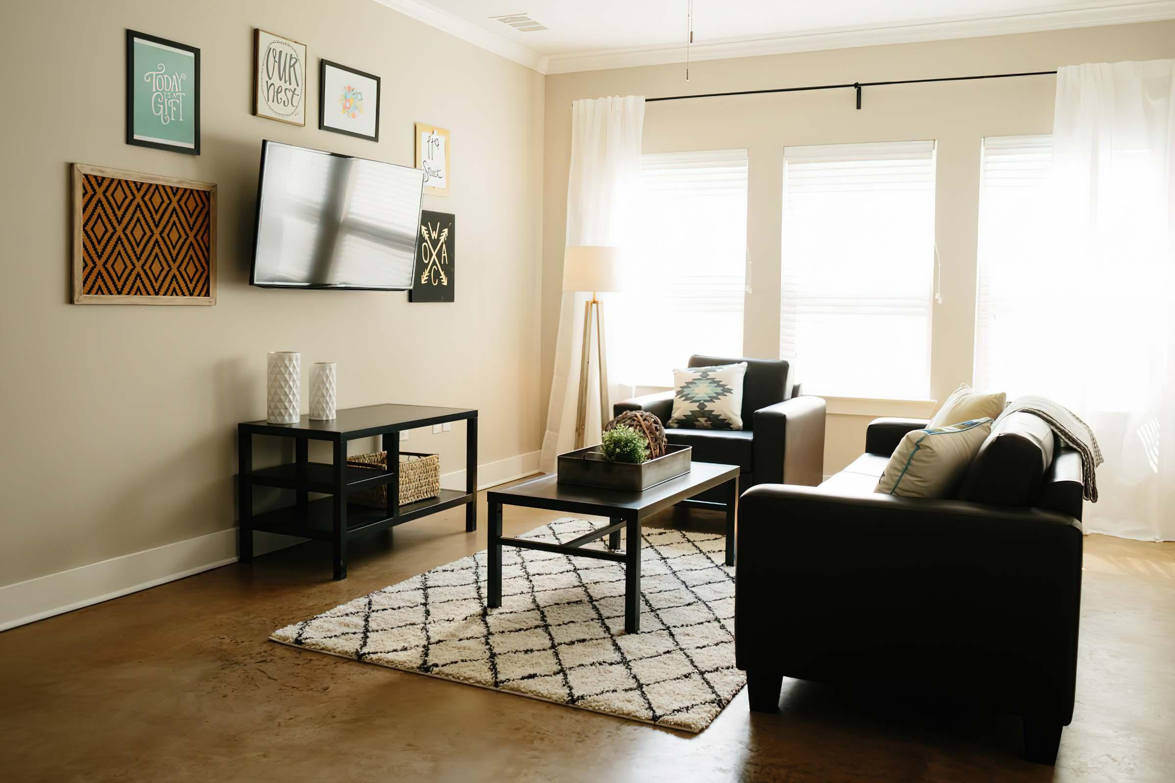 A cozy living room featuring a black leather sofa and armchair, a black coffee table, and a TV mounted on the wall. The room has light-colored walls adorned with framed artwork and two windows with sheer curtains. A patterned rug covers the floor, and a tall lamp adds a warm touch to the space.
