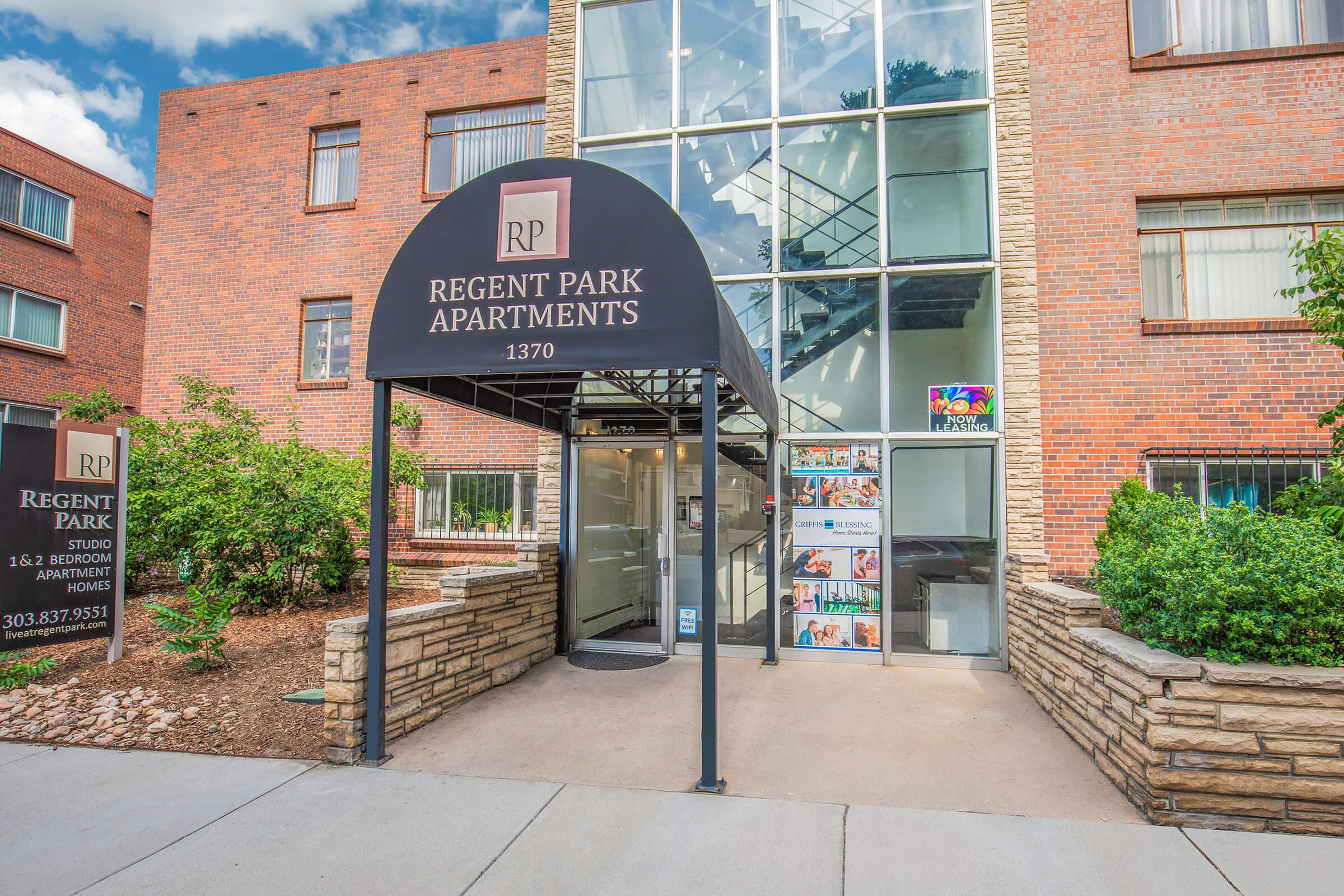 Entrance of Regent Park Apartments, featuring a black awning with the logo. The building has a mix of brick and glass architecture. On the left, there's a landscaped area with rocks and greenery. Nearby, a sign displays contact information. The overall atmosphere is welcoming and well-maintained.