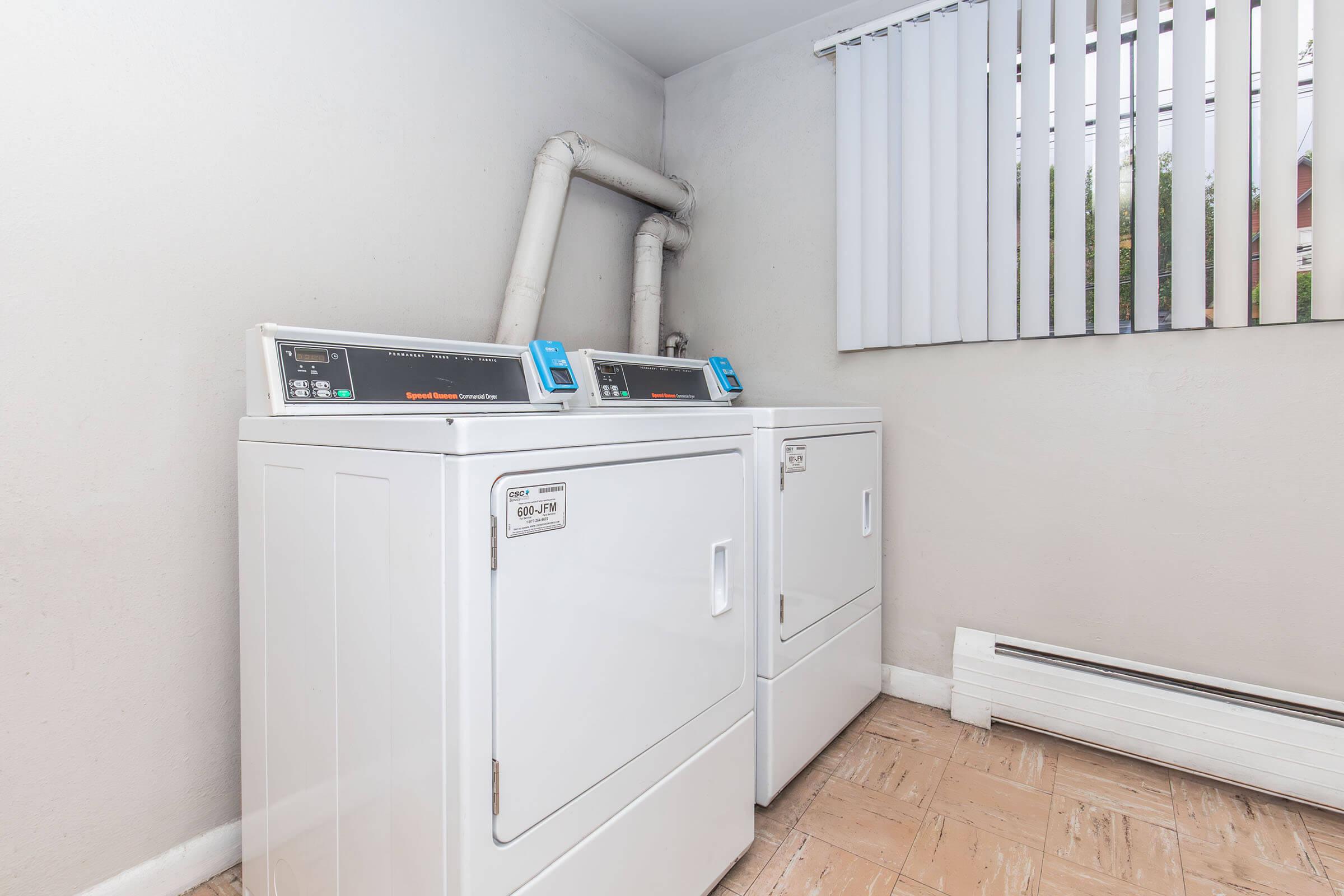 Two stacked white laundry machines are positioned in a small, well-lit laundry room. The machines are modern and feature digital displays. A window with white blinds allows natural light to fill the space, which has tiled flooring and neutral-colored walls.