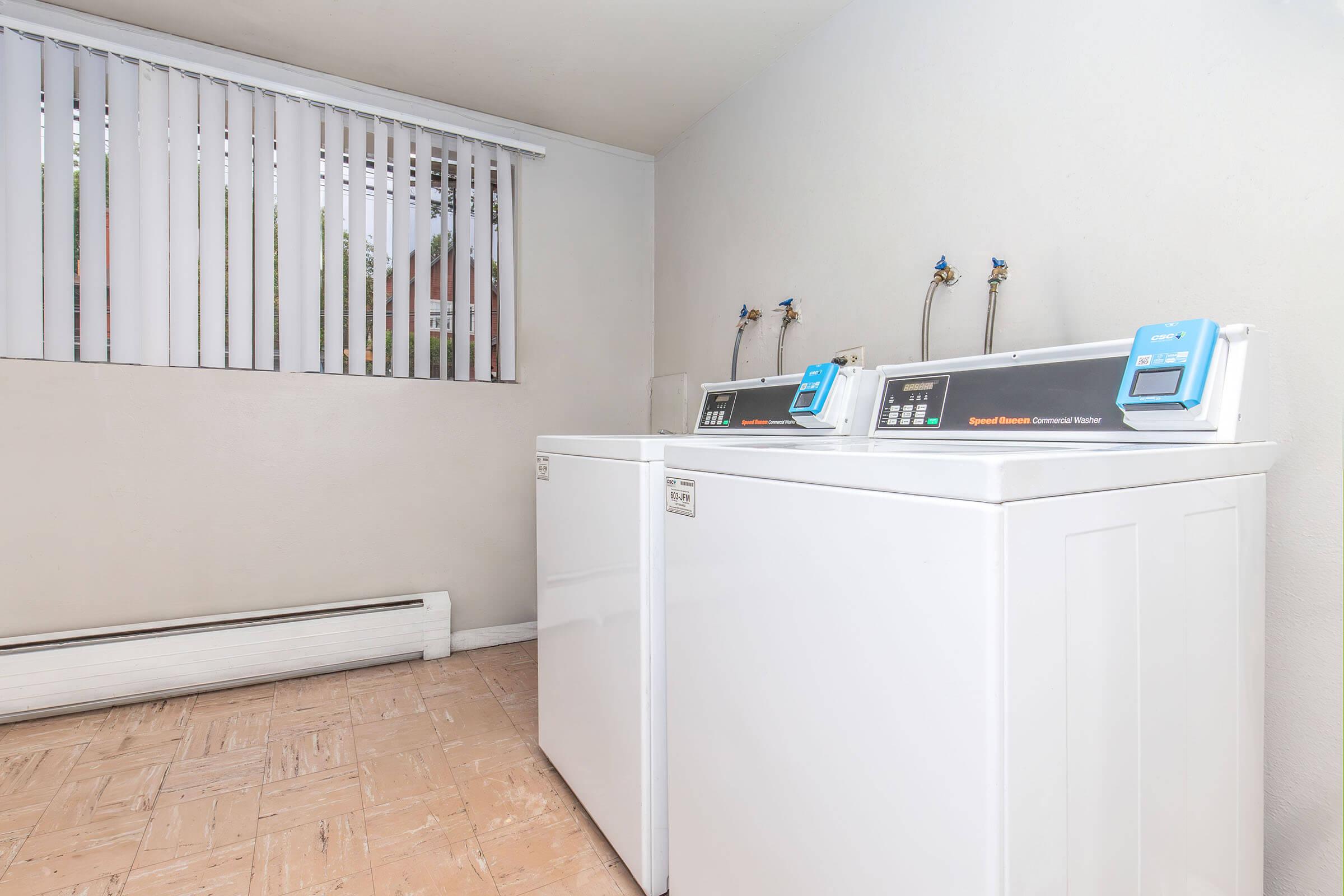 A clean laundry room featuring two white commercial washing machines against a light-colored wall. There are window blinds providing natural light and a radiator along the floor. The space appears organized and well-maintained, suitable for laundry activities.