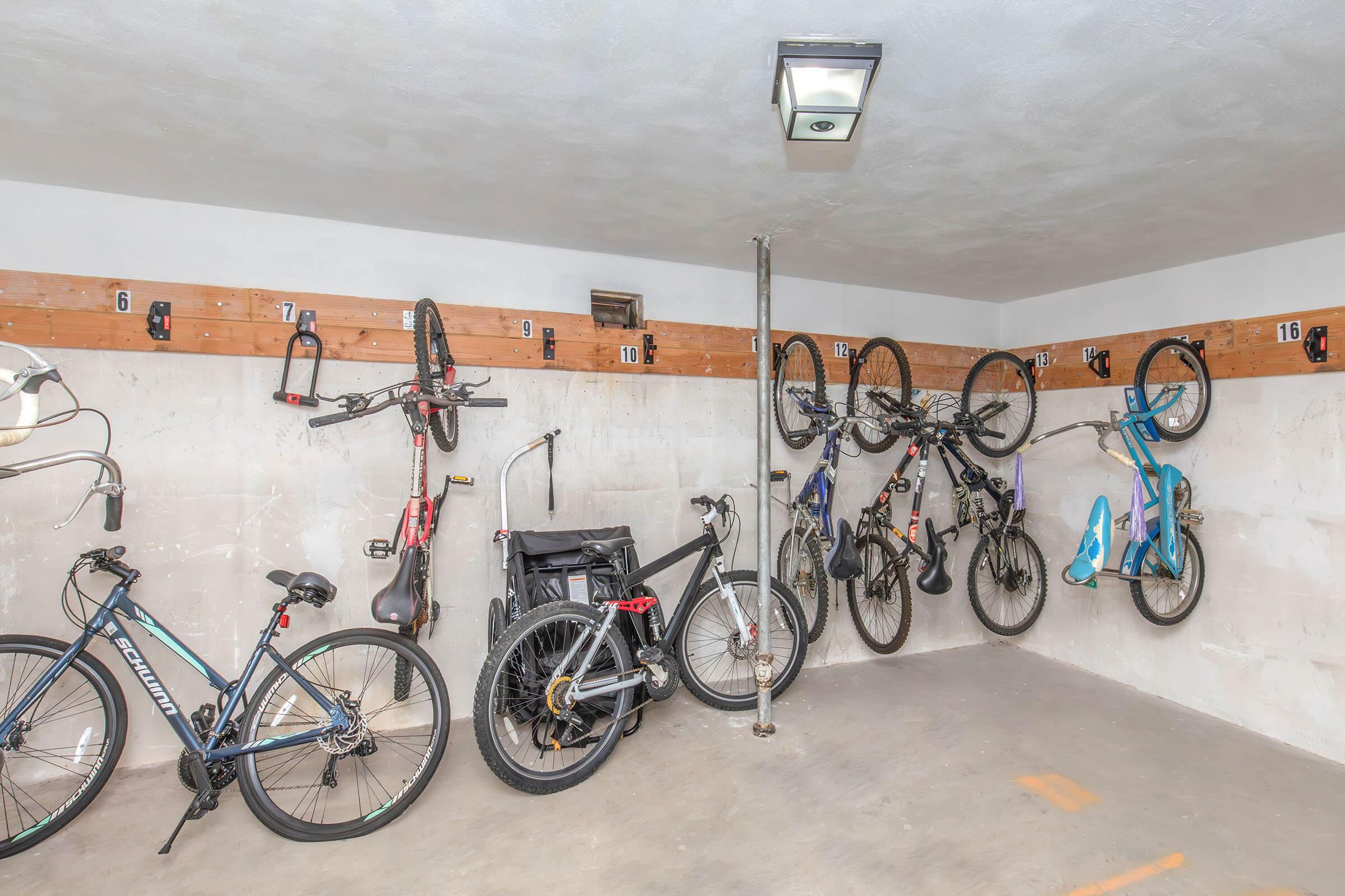 A well-organized garage space featuring several bicycles of various styles. The bicycles are hanging from wall-mounted hooks, with some resting against a concrete wall. A wall-mounted light fixture illuminates the area, and the floor is bare concrete.