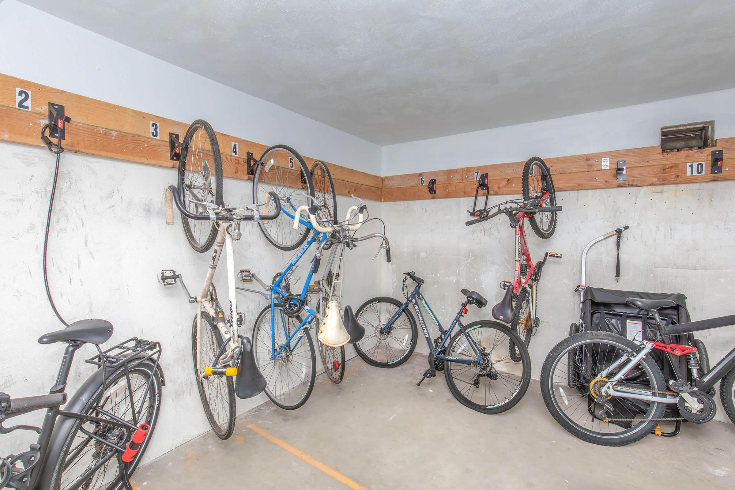 A garage or storage area featuring multiple bicycles hanging on wall-mounted hooks. The bikes vary in color and style, with a couple of them being mountain bikes and a road bike. The walls are plain and the floor is concrete, suggesting a functional space for bike storage.