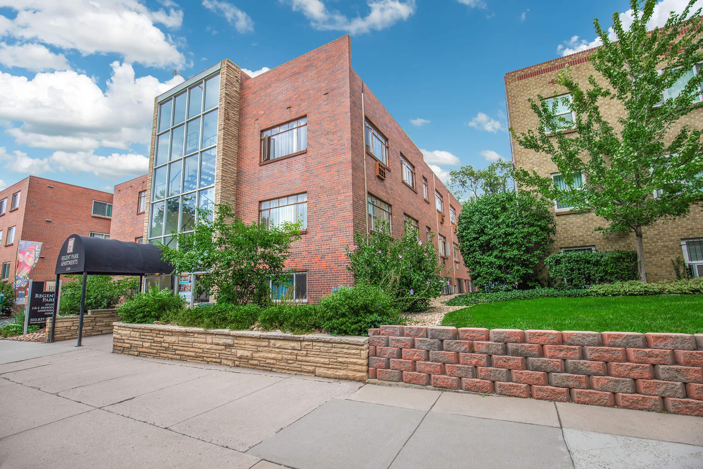 A modern brick apartment building with large windows, surrounded by greenery and well-maintained landscaping. The exterior features a mix of brick and stone, and a canopy entrance. The sky is partly cloudy, creating a bright and inviting atmosphere.