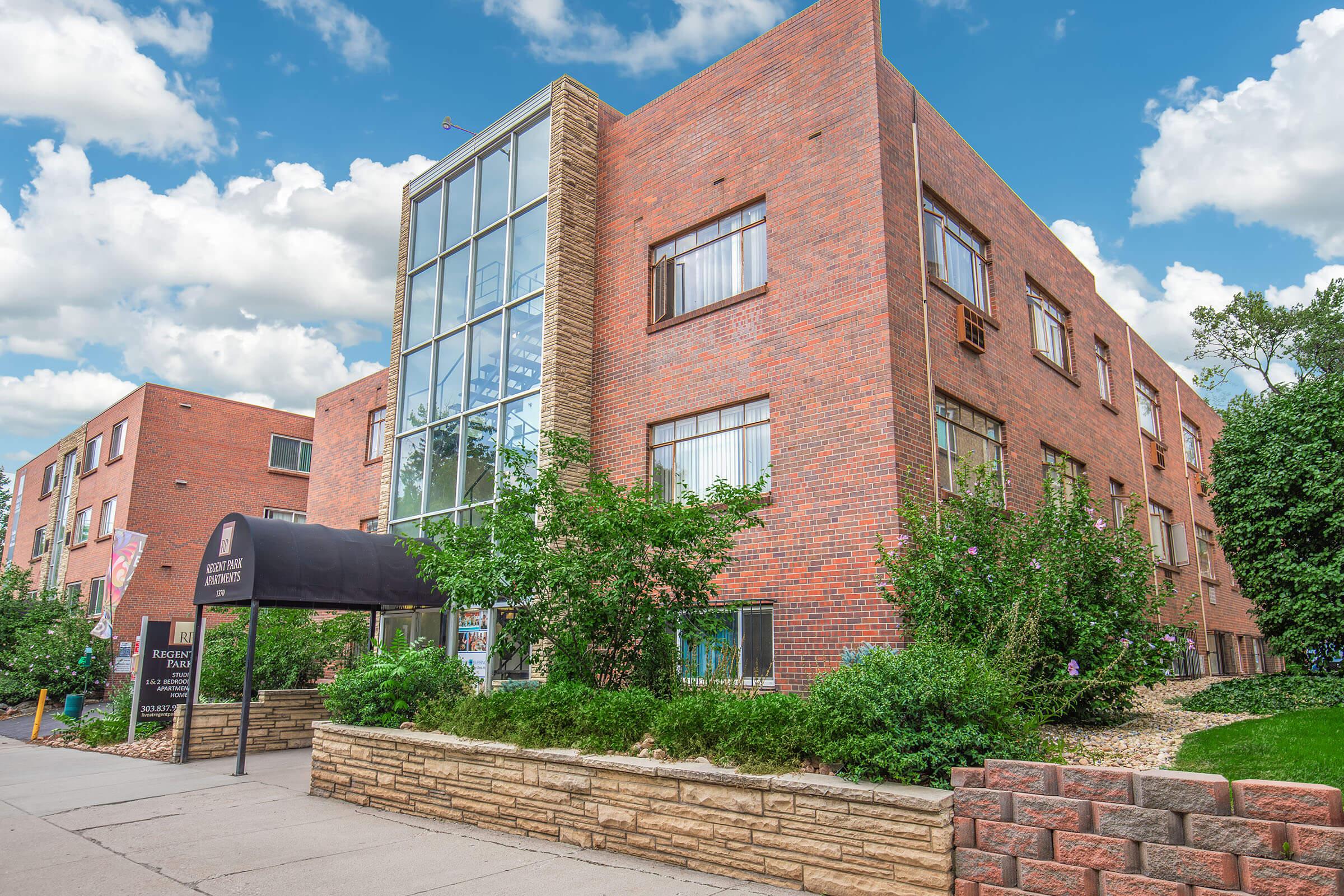A modern red brick building featuring large windows, a glass entrance, and landscaped greenery in front, under a blue sky with fluffy clouds. The structure has multiple floors and an awning at the entrance, indicating it may be a residential or commercial property.