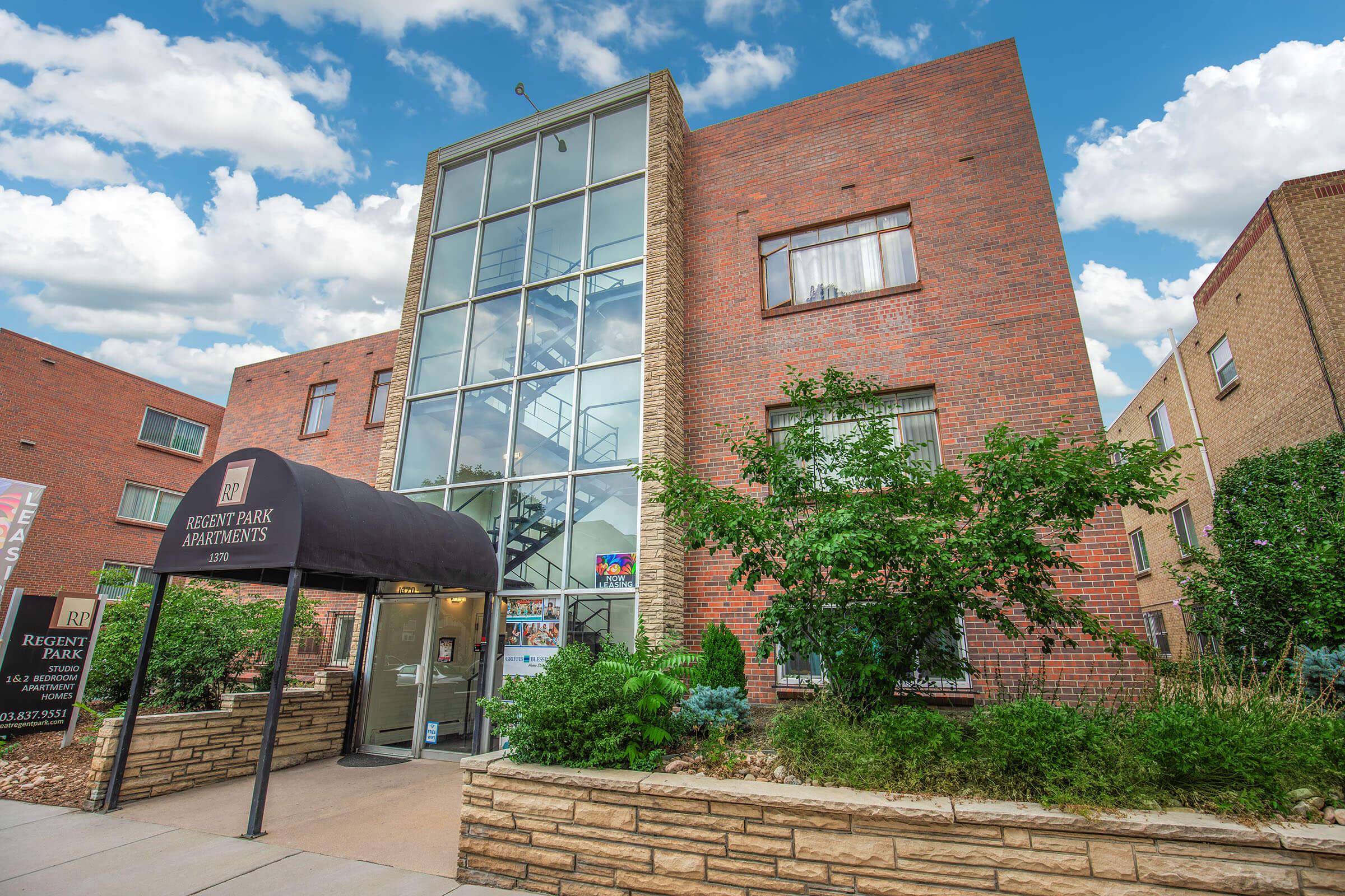A multistory brick building with large windows, featuring a black awning at the entrance labeled "Regent Park Apartments." Lush green shrubs and trees surround the entrance, and a blue sky with fluffy clouds is visible in the background. The overall setting appears welcoming and well-maintained.