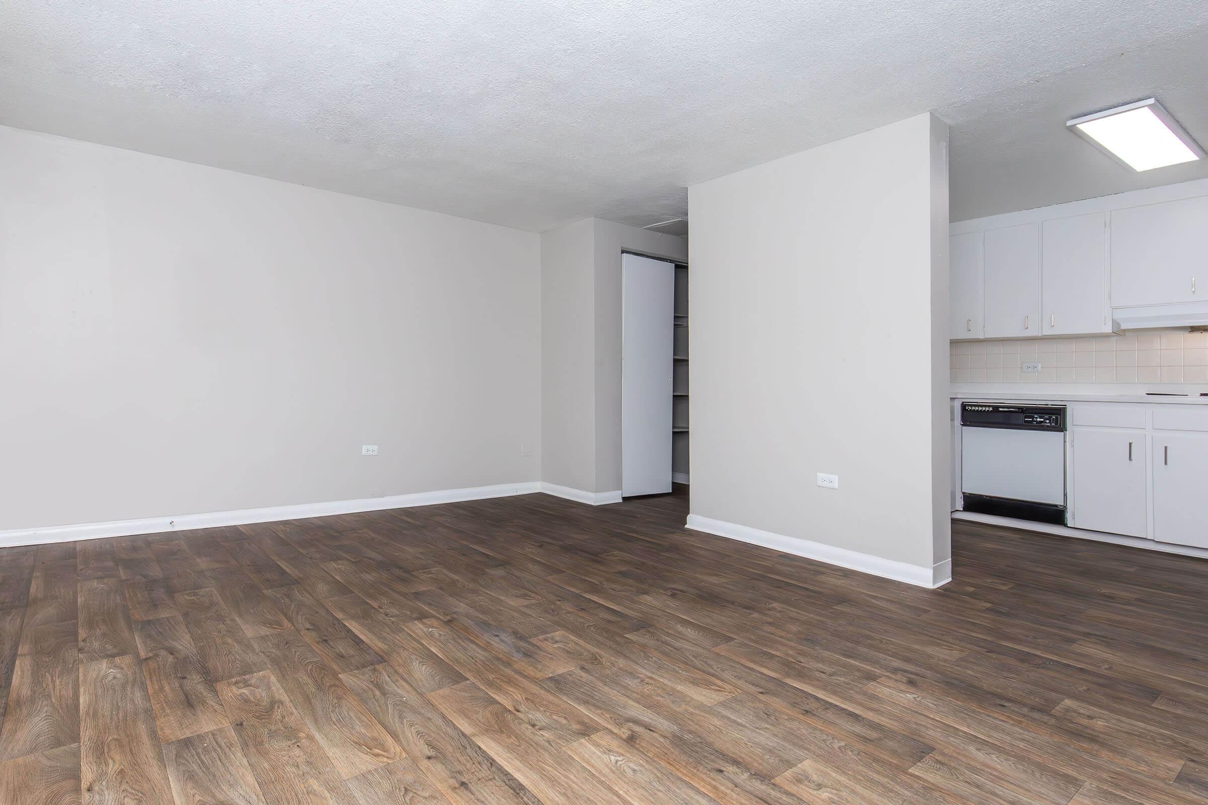 A spacious, empty room with light-colored walls and a modern wood-like floor. On one side, there is a small kitchen area visible with white cabinets, a stove, and a dishwasher. The room has ample natural light, giving it a bright and airy feel.
