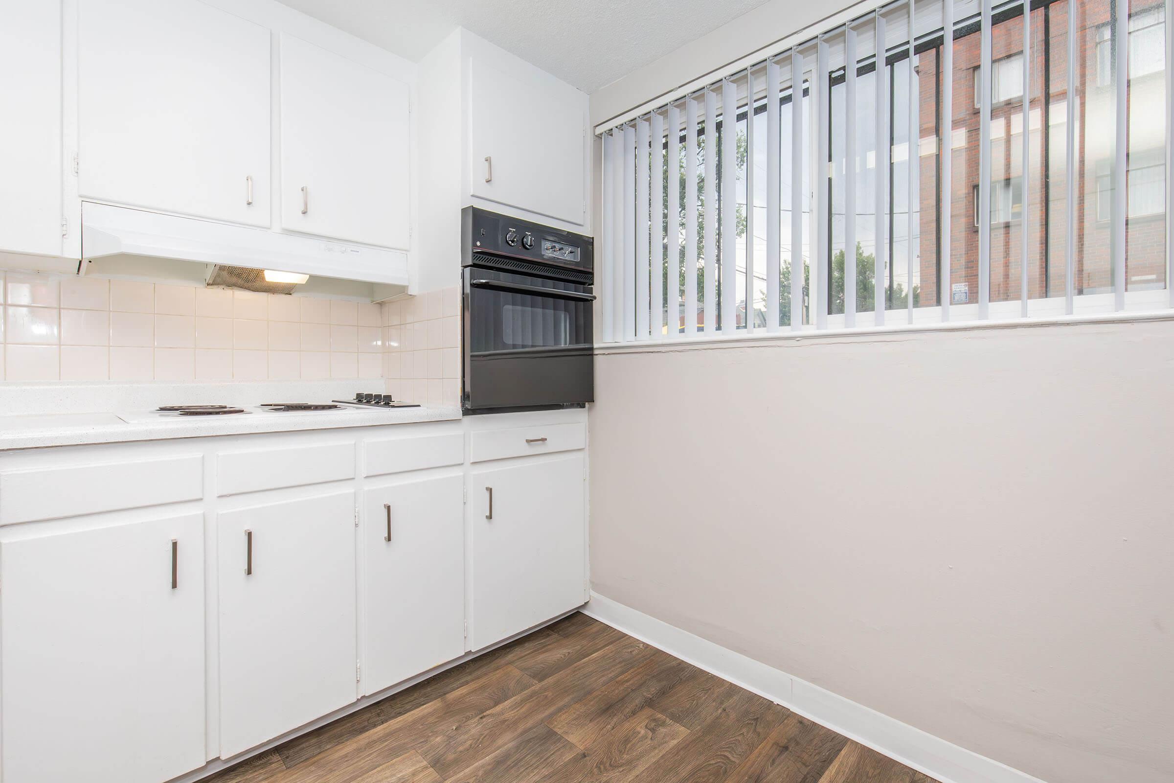 Bright, modern kitchen with white cabinets, a black oven and stove, and a window with vertical blinds. The flooring is a warm wood tone, adding a cozy feel to the space. The kitchen is clean and uncluttered, creating an inviting atmosphere.