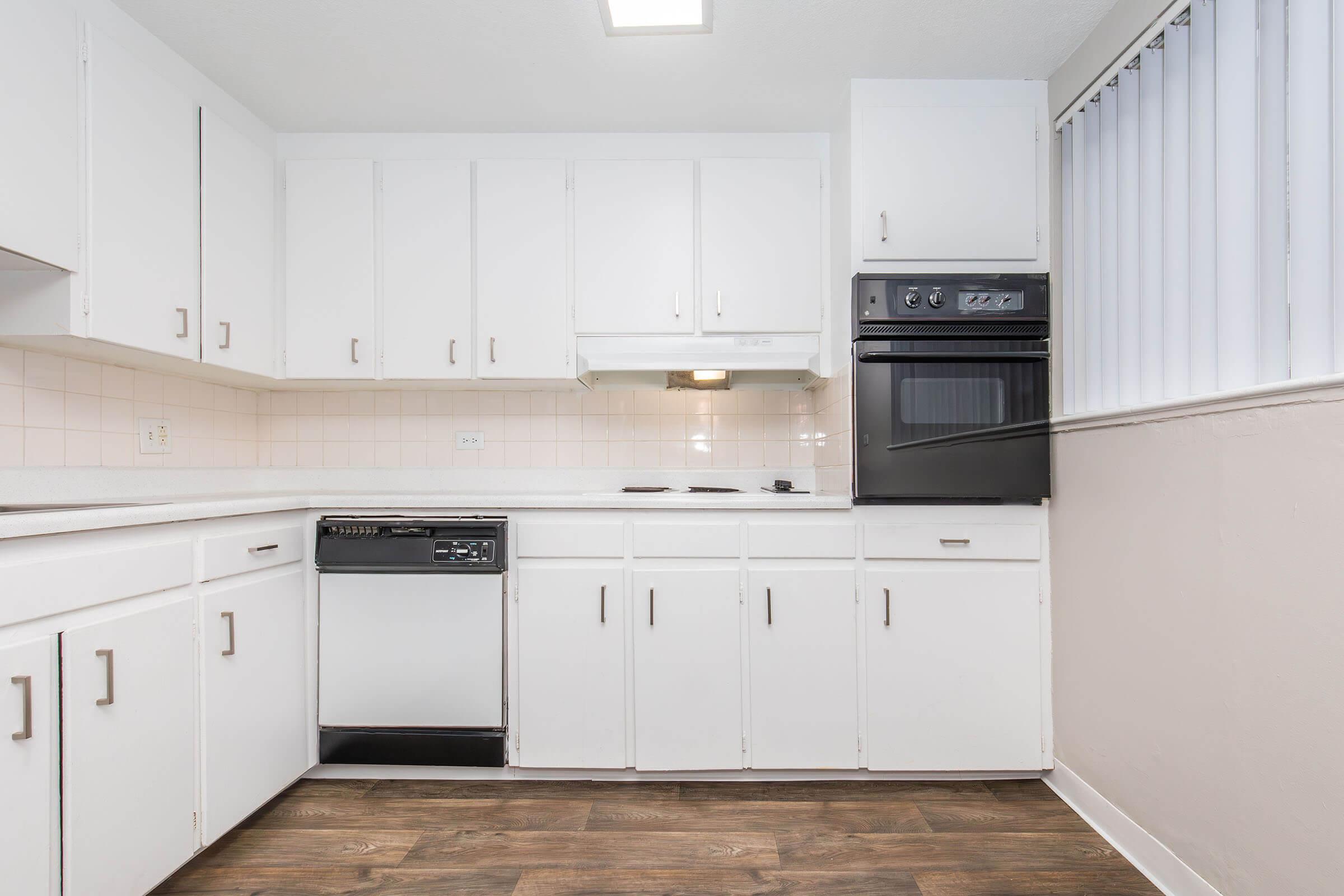 Bright kitchen featuring white cabinets, a black oven and range, a dishwasher, and a countertop. The room has a window with vertical blinds and a light-colored tiled backsplash. The flooring is a wood-look laminate, giving a modern and clean aesthetic.