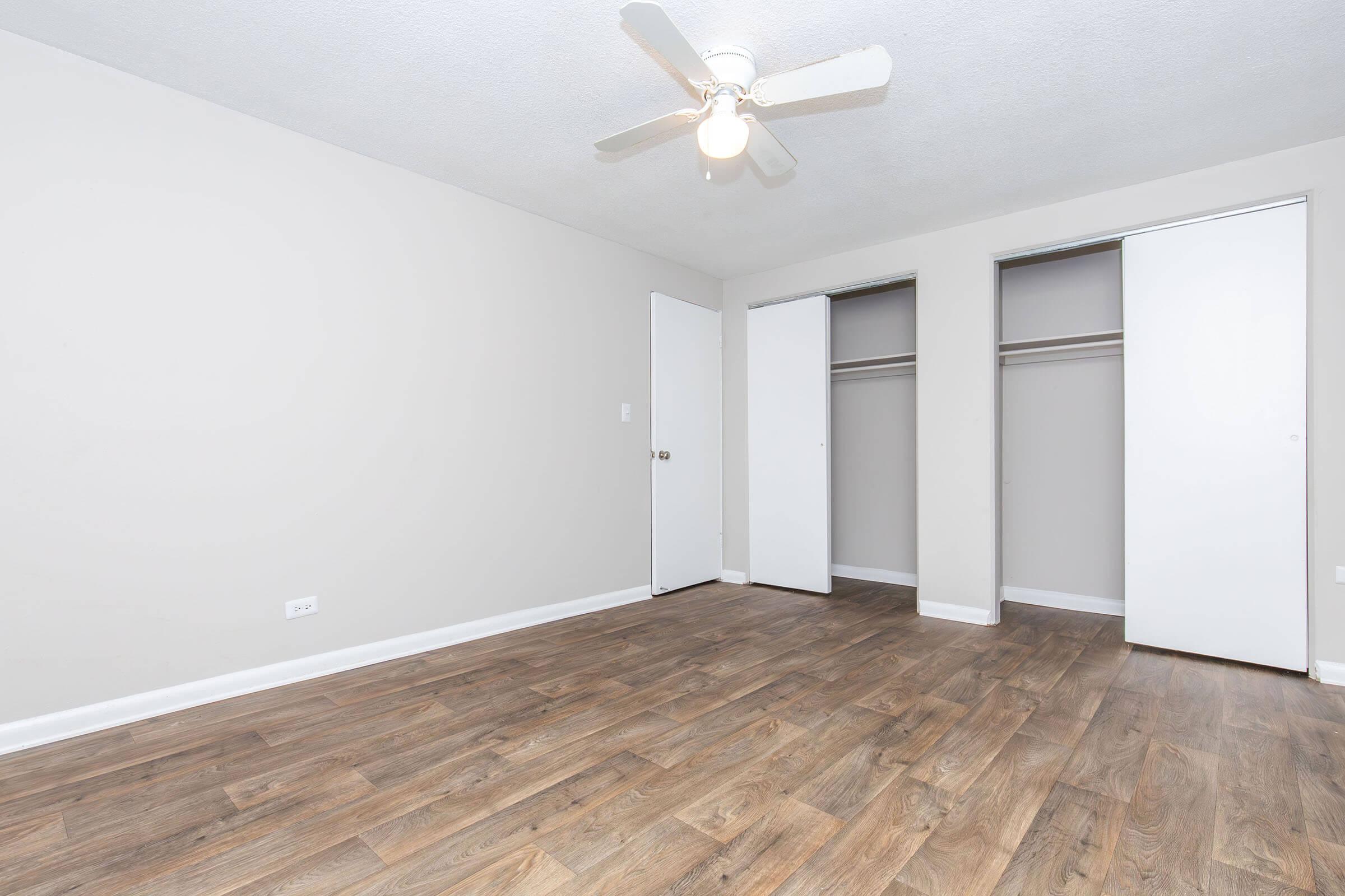 A vacant room featuring light-colored walls, wooden flooring, and a ceiling fan. There are two white closet doors on the right side, and a closed door is visible on the left. The space is well-lit and lacks furniture, creating an open and airy atmosphere.