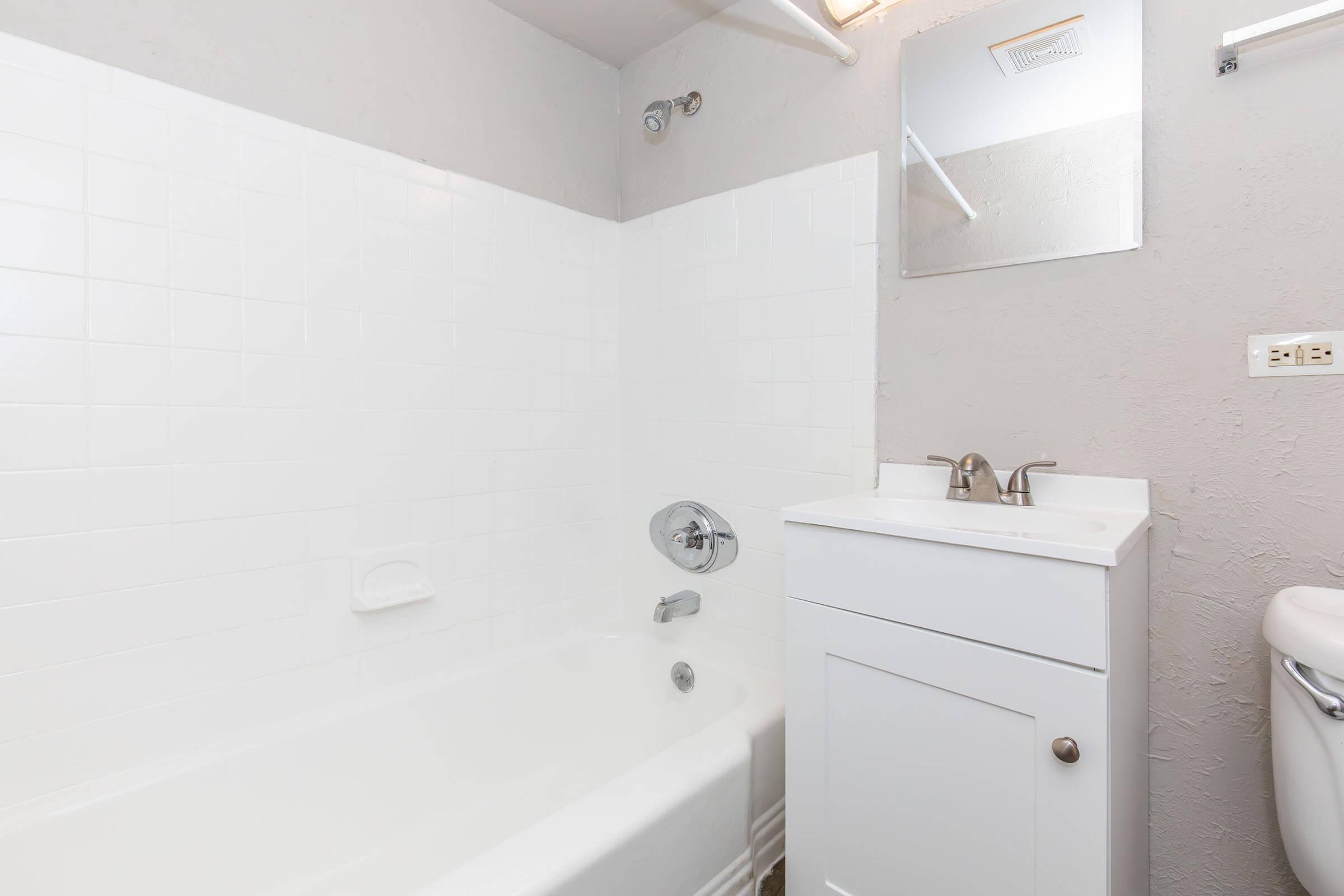 A clean, modern bathroom featuring a white tub with a showerhead, a small sink vanity with two faucets, and a mirror above the sink. The walls are light gray, and the floor is not visible in the image. A toilet is in the corner, completing the simple yet functional layout.