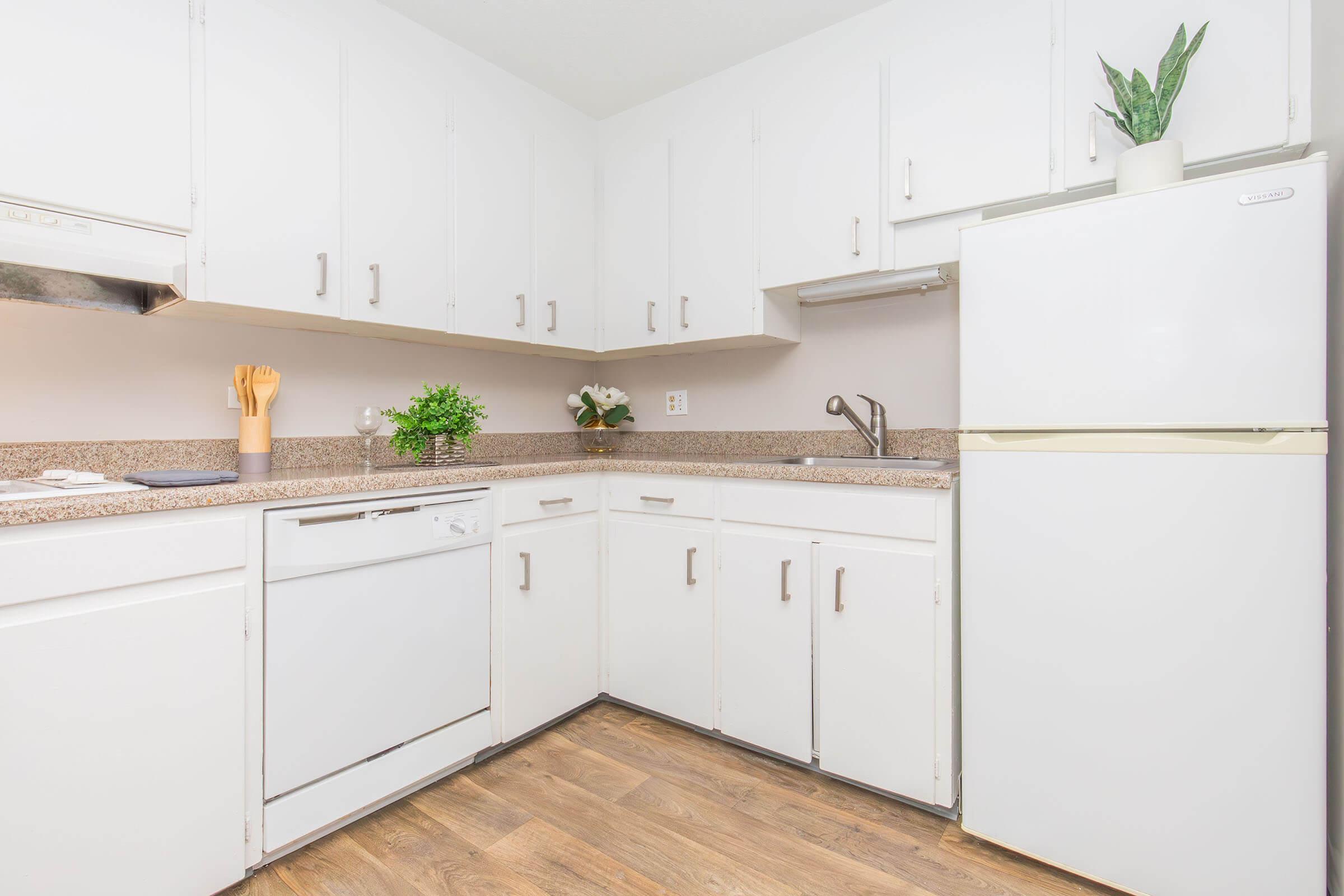 A clean, modern kitchen featuring white cabinets, a granite countertop, a stainless steel dishwasher, and a white refrigerator. The space is adorned with a potted plant and decorative items, with wooden flooring that adds warmth to the overall design. Natural light brightens the room.
