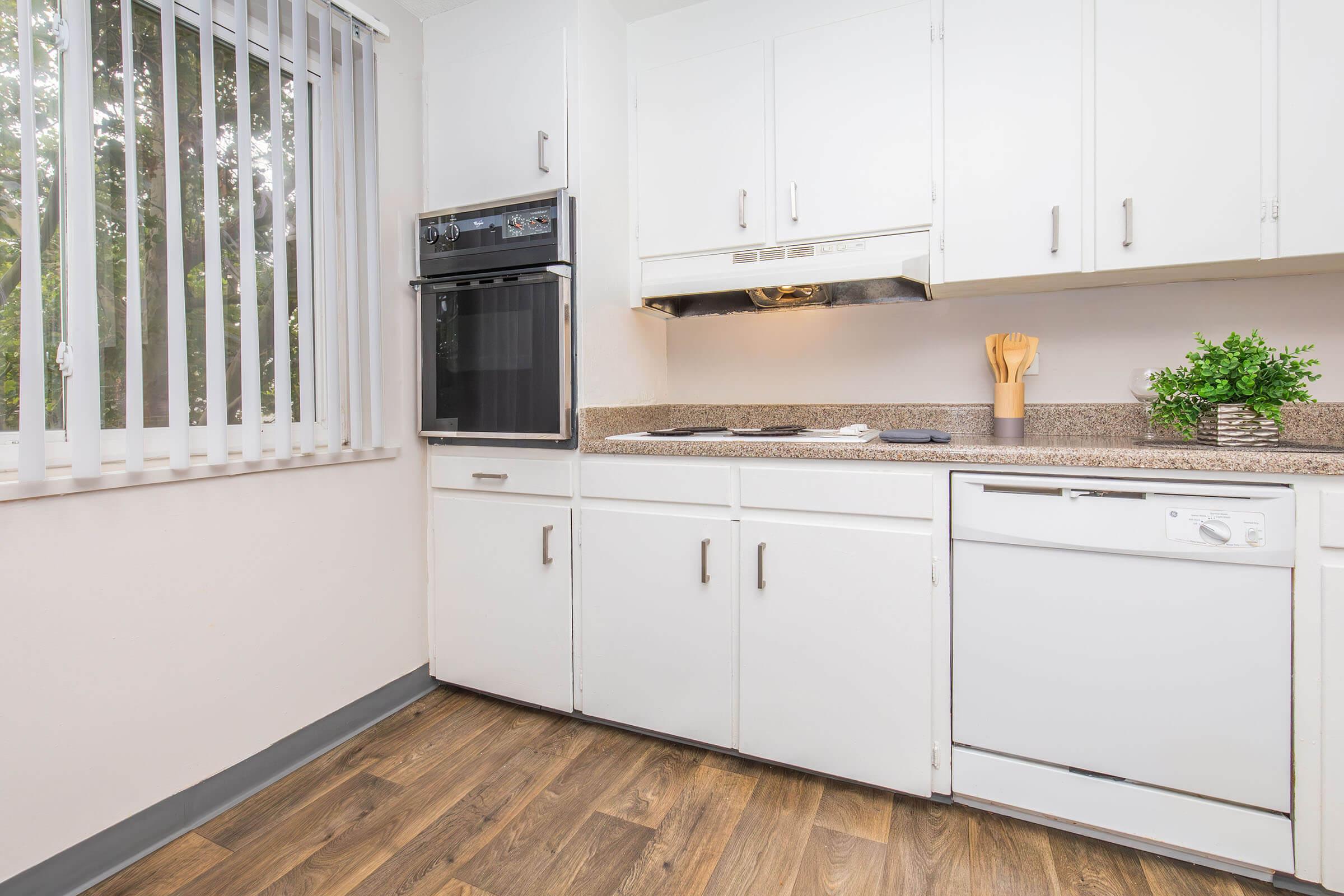 Modern kitchen with white cabinetry, a countertop, and built-in appliances including an oven and dishwasher. Natural light filters through a window with vertical blinds, and there's a small green plant on the counter, adding a touch of greenery to the space. Wood-style flooring completes the look.