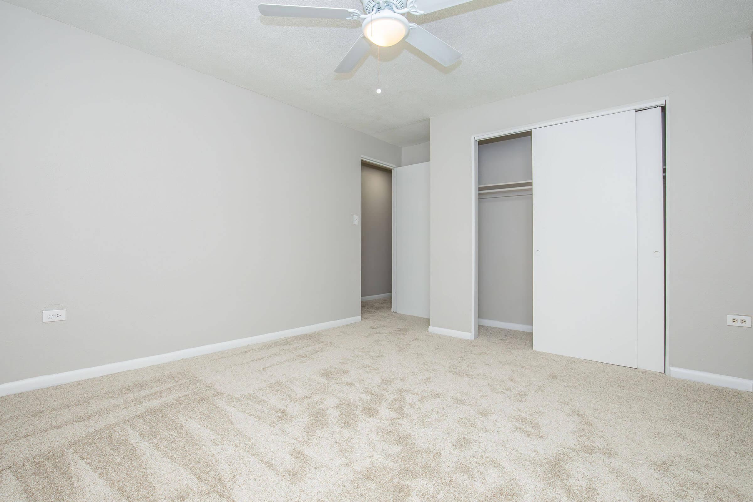 A spacious, empty bedroom with light gray walls and a ceiling fan. The floor is covered in beige carpet. There are two doorways, one leading to a closet with sliding doors. Natural light enters through an unseen window, creating a bright and airy feel.