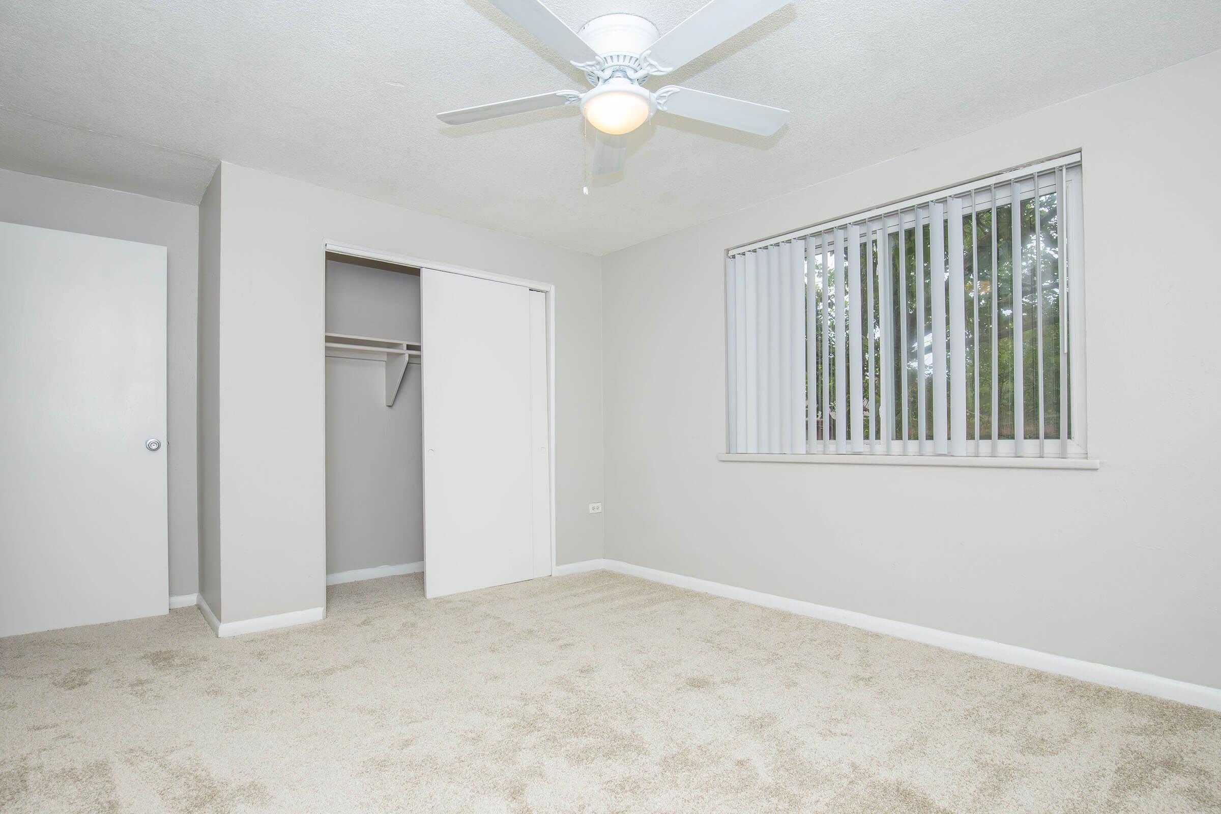 A spacious, vacant bedroom featuring light gray walls, a ceiling fan, and large windows with vertical blinds. The room has beige carpet flooring and an open closet with sliding doors, providing a clean and modern look. Ideal for decorating and personalizing.