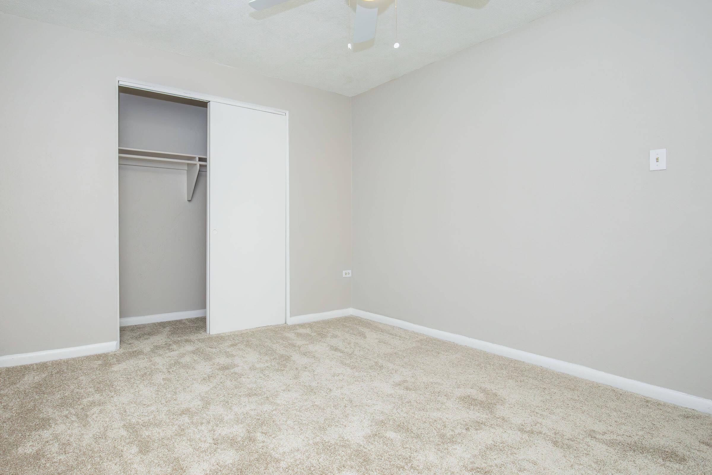 A clean, empty room with light gray walls and a ceiling fan. The floor is covered in beige carpet. To the left, there is a white closet door partially open, revealing a simple closet interior. The room is well-lit, creating an airy and spacious feel.