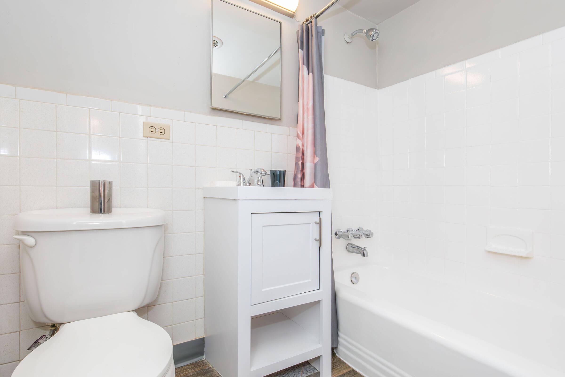 A clean bathroom featuring a white toilet, a small vanity with a sink, and a bathtub with a shower. The walls are tiled in white and light gray, and there is a mirror above the sink. A shower curtain in a soft color adds a touch of decor. The floor is wood-like, adding warmth to the space.