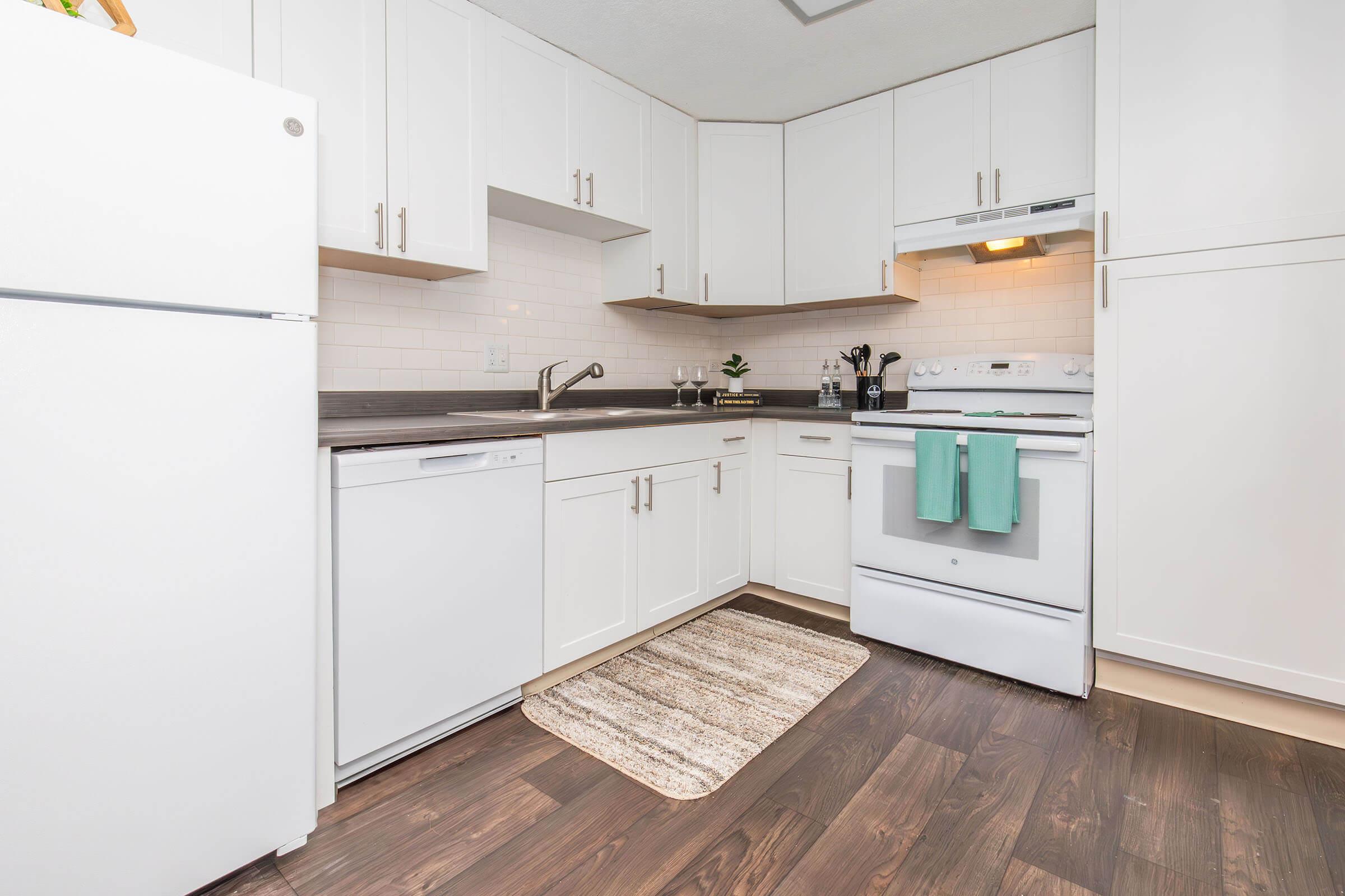 Modern kitchen featuring white cabinets, a stainless steel refrigerator, a dishwasher, and a white stove with an overhead range hood. The countertop is dark wood, and a small plant decorates the space. A striped mat lies on the floor, and a light fixture casts warm light over the area.