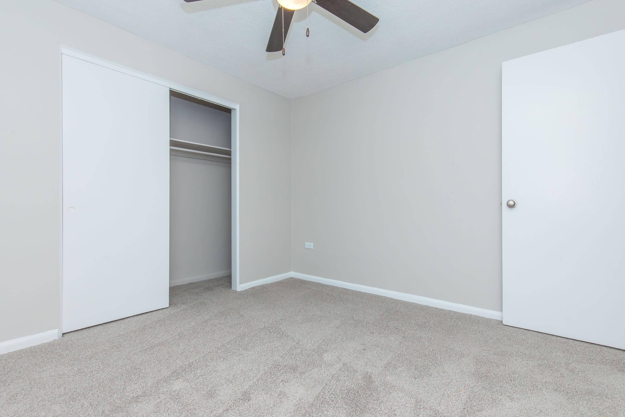 A simple, empty room with light gray walls and beige carpet. There are two white doors: one leading to a closet on the left and a regular door on the right. A ceiling fan is mounted above, and the space is well-lit with natural light.