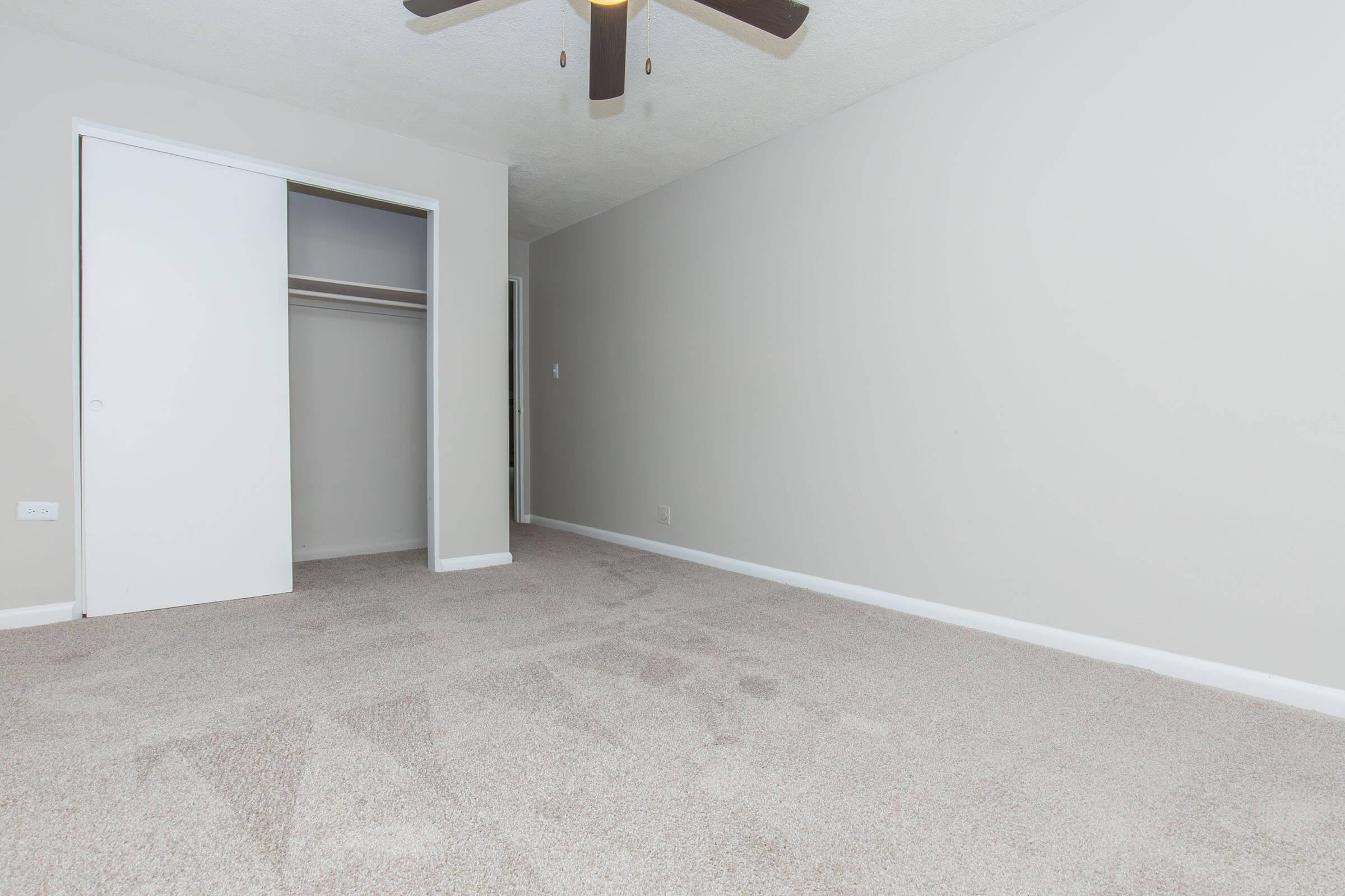 A spacious, empty room with light gray walls and plush beige carpet. There is a ceiling fan with wooden blades, a white closet door open to reveal a closet space, and another door leading to a hallway. The room is well-lit, creating a clean and inviting atmosphere.