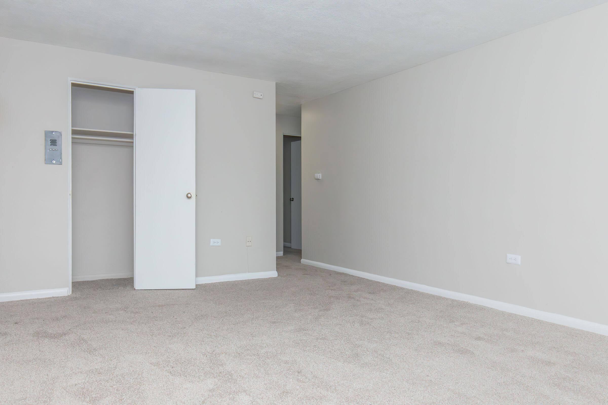 A spacious, empty room with light beige walls and a soft carpeted floor. There is a white door leading to a closet on the left, and a door frame visible on the right side, suggesting access to another room or hallway. Natural light may be coming from an unseen window.