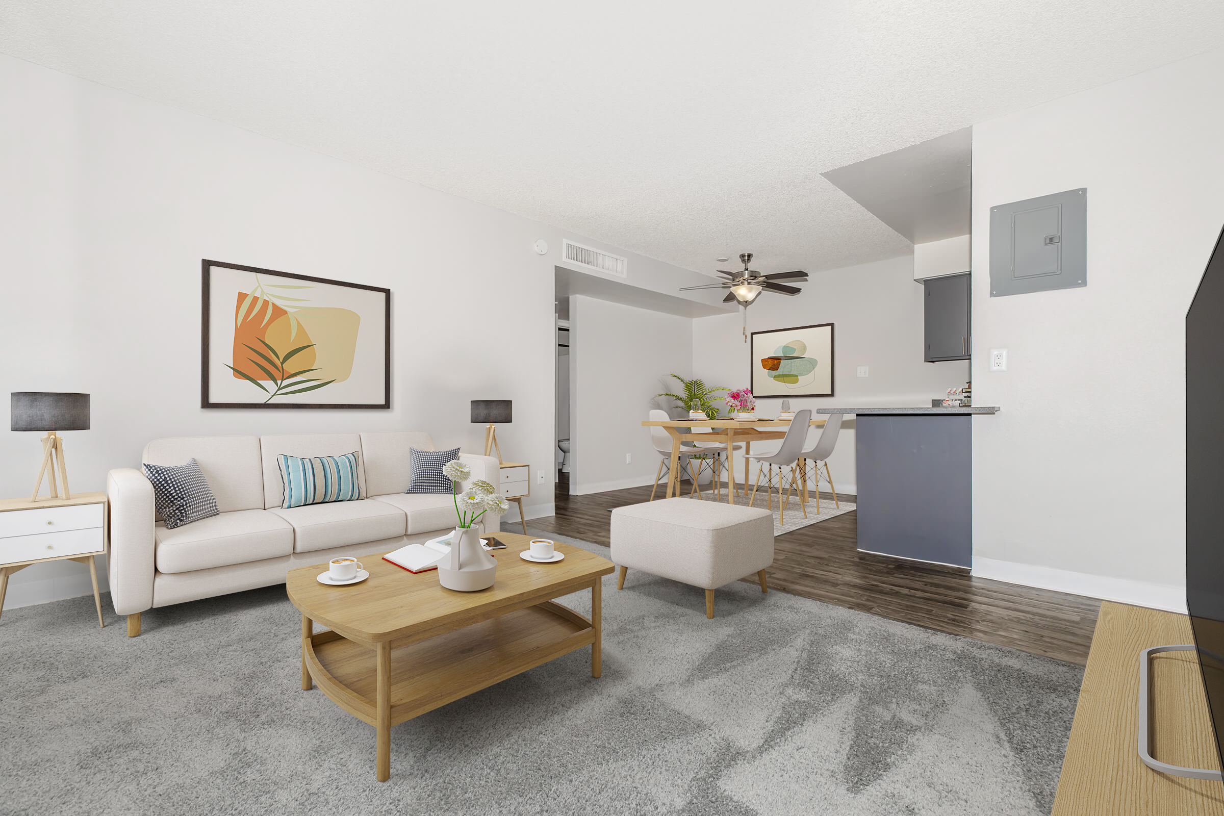 Bright and modern living room featuring a light-colored sofa with decorative pillows, a wooden coffee table with cups, and a plush area rug. The walls have stylish artwork, and there are plants for decoration. A dining area with a table and chairs is visible in the background, along with a ceiling fan and kitchen area.