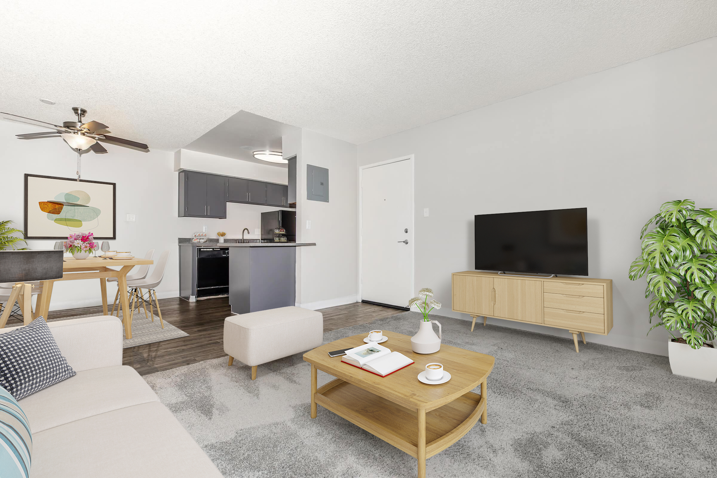 A modern living room featuring a light-colored sofa with decorative pillows, a wooden coffee table with books and a mug, and a flat-screen TV on a wooden media console. The space includes a dining area with a small table and chairs, and a potted plant in the corner, all set in a bright, airy environment.