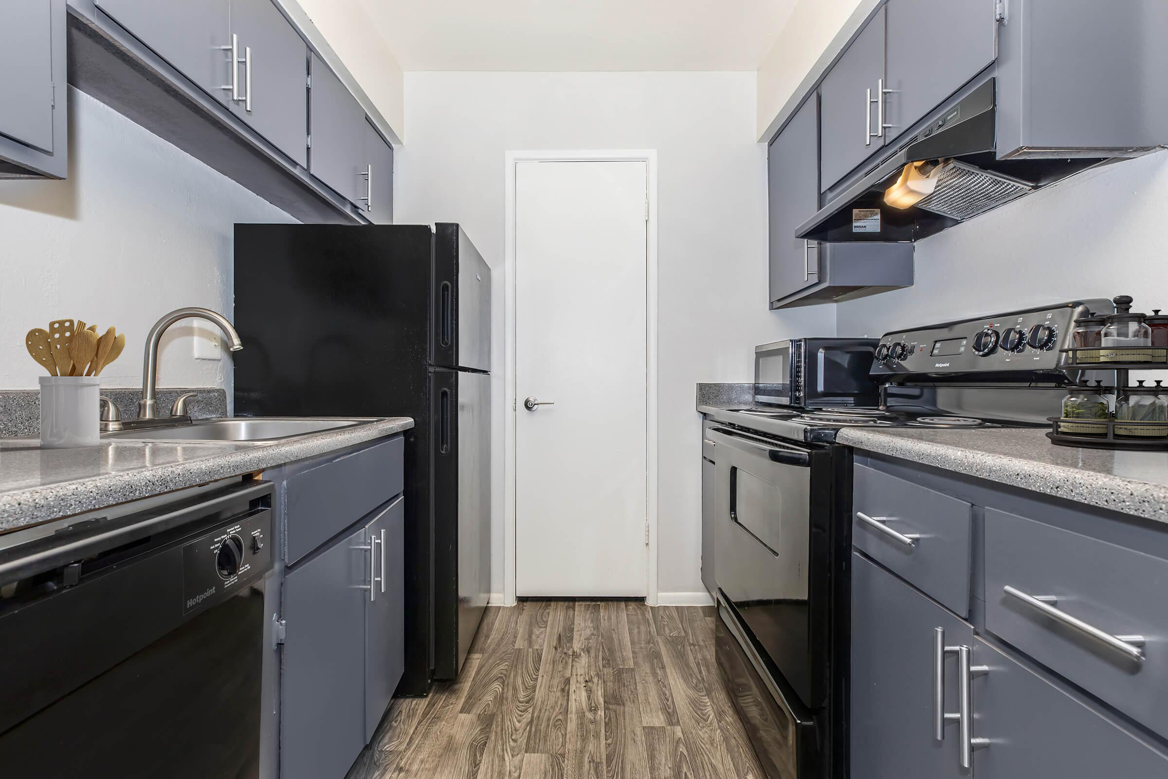 A modern kitchen featuring grey cabinets and countertops. The space includes a stainless steel sink, black appliances such as a refrigerator, microwave, and oven. The floor is a light wood laminate, creating a sleek and contemporary look. A door leads to another room, adding depth to the layout.