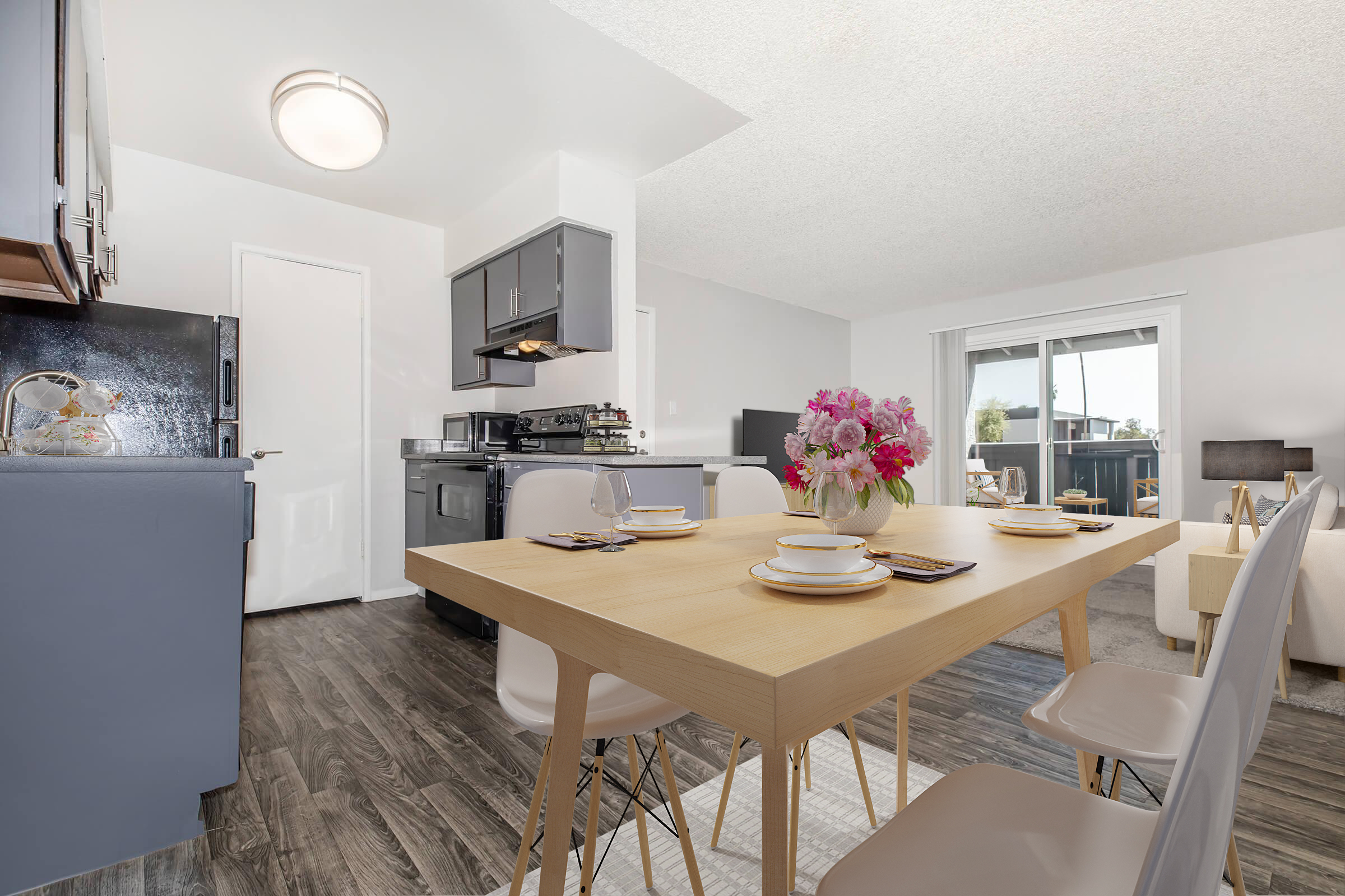 Bright, modern kitchen and dining area featuring a wooden table set for dinner with plates and utensils. White walls, stylish chairs, and a bouquet of flowers as a centerpiece. Kitchen appliances visible, with a view of a balcony and outdoor light coming through large windows. Cozy and inviting atmosphere.