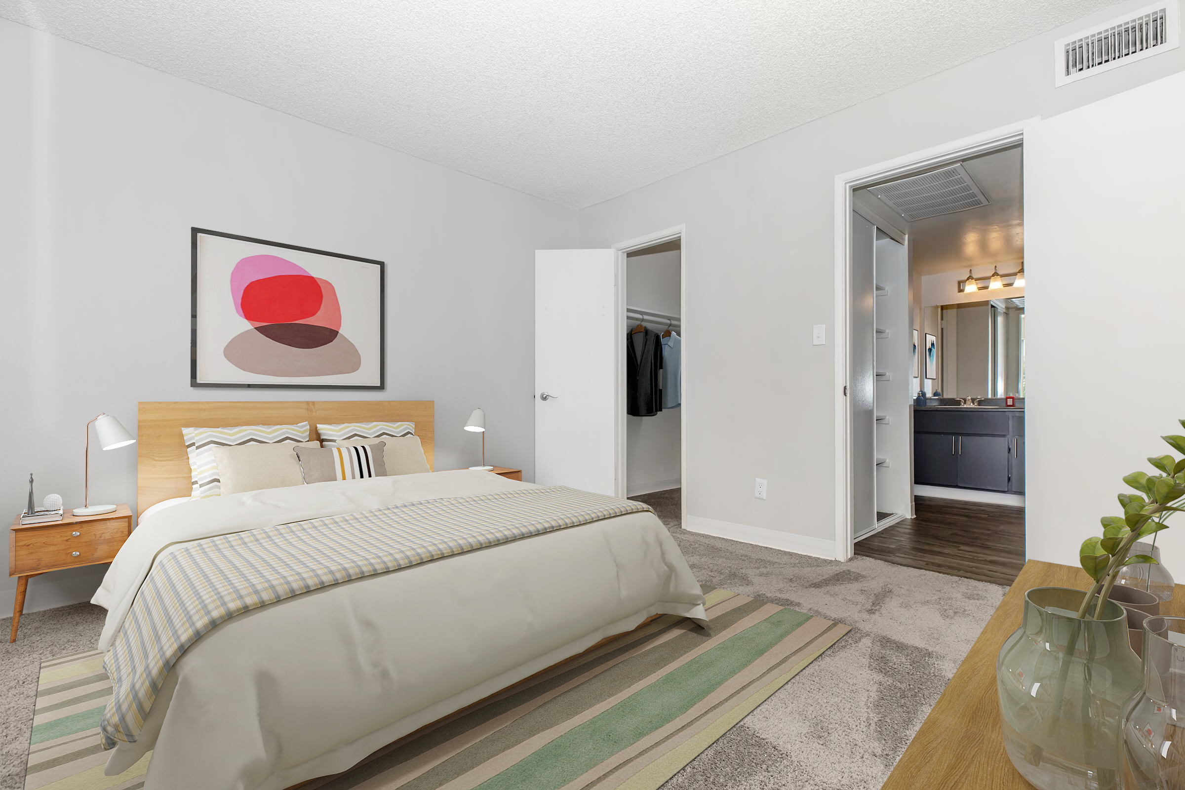 A modern bedroom featuring a wooden bed frame with a cream-colored duvet and decorative pillows. There are two bedside lamps on nightstands, a cozy rug on the floor, and a closet with hanging clothes. In the background, a bathroom with a double vanity and mirror is visible. The walls are painted light gray.