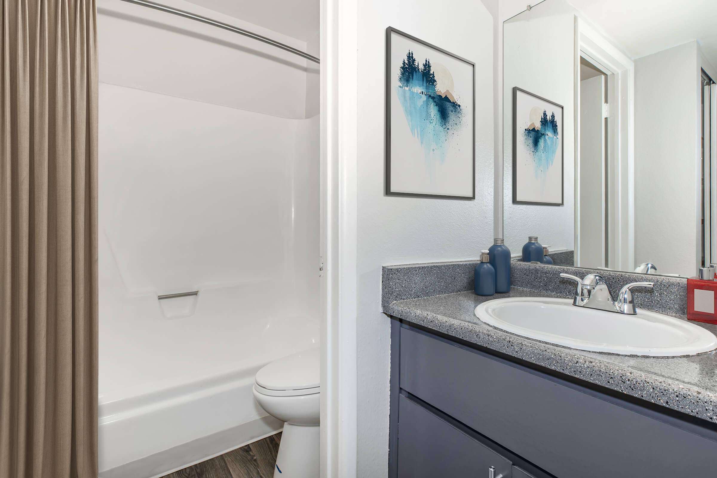 A modern bathroom featuring a shower stall with a curtain, a toilet, and a sink with a gray countertop. Two framed art pieces with blue abstract designs hang on the wall. The cabinetry is dark gray, and there are two decorative items on the countertop. The overall decor is clean and contemporary.