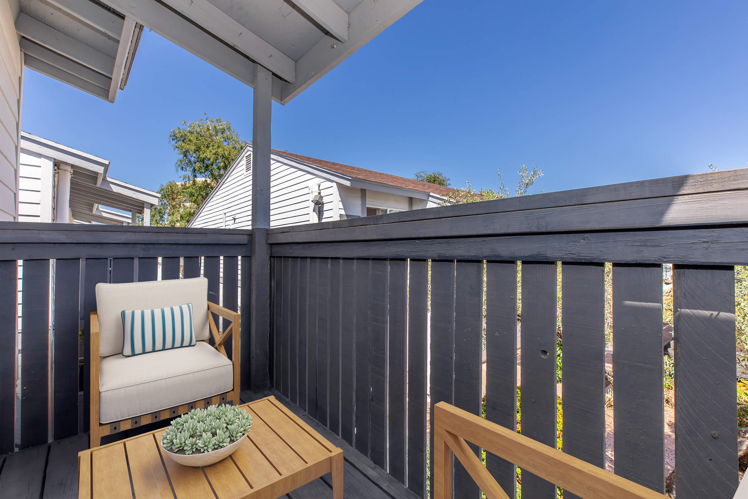 A cozy outdoor seating area featuring a light-colored armchair with blue-striped cushions and a wooden table. A small potted succulent sits on the table, surrounded by a wooden railing and a clear blue sky above, creating a serene backyard scene.