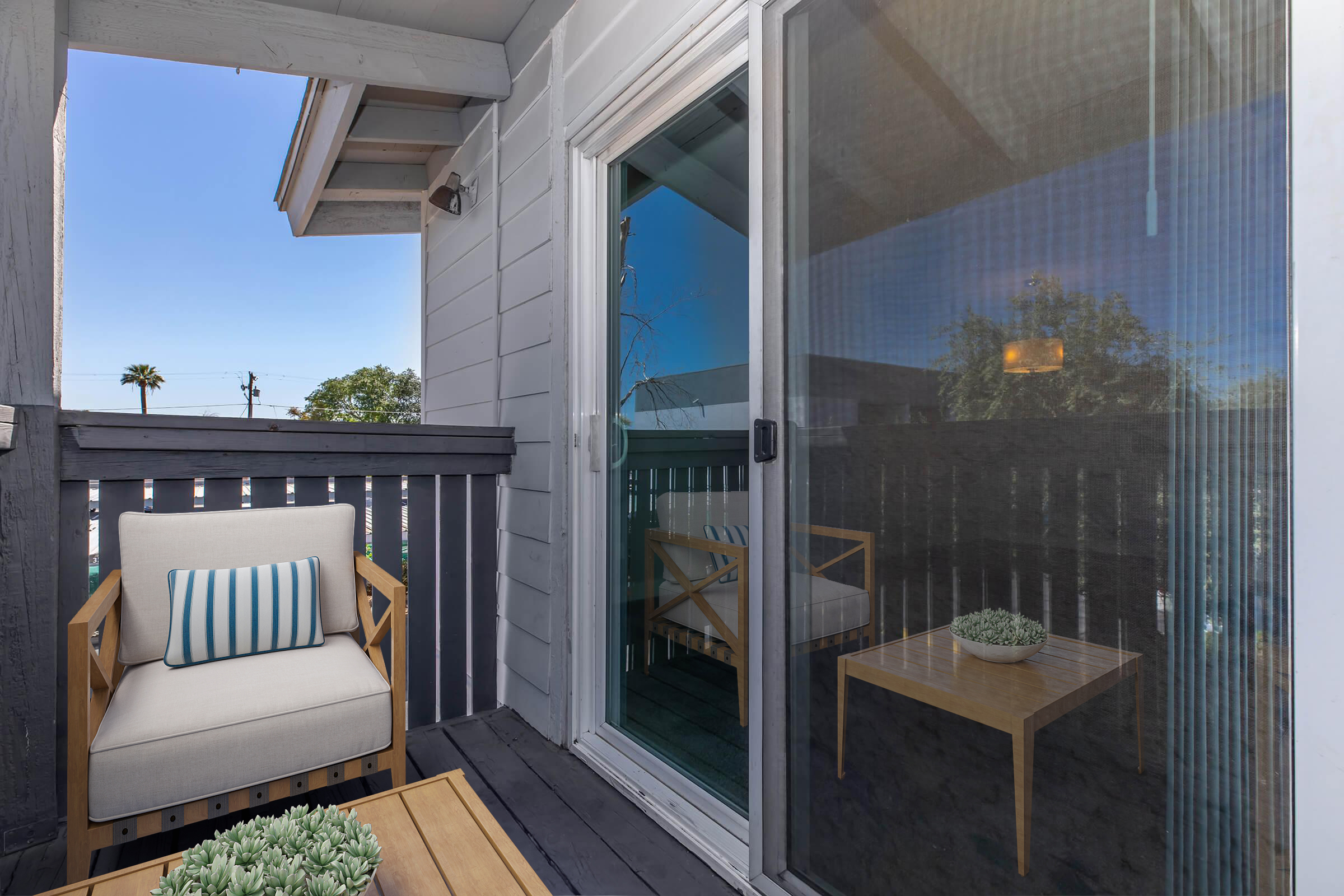 A small balcony featuring a light-colored armchair with a striped cushion and a wooden table. The scene includes a sliding glass door, a clear blue sky, and a few green plants visible in the background. The balcony railing is painted dark, creating a cozy outdoor space.