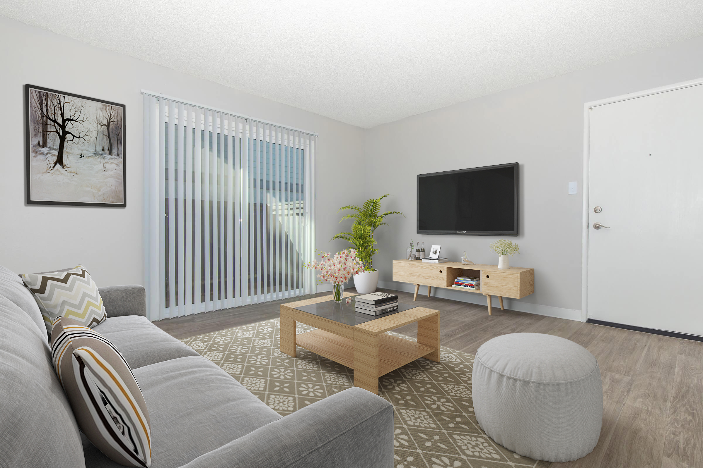 A modern living room featuring a light gray sofa, a wooden coffee table with books and flowers, and a round ottoman. A television mounted on the wall is opposite the sofa. Large sliding glass doors with vertical blinds lead to an outdoor space, and a wall art piece depicting a winter scene is displayed.