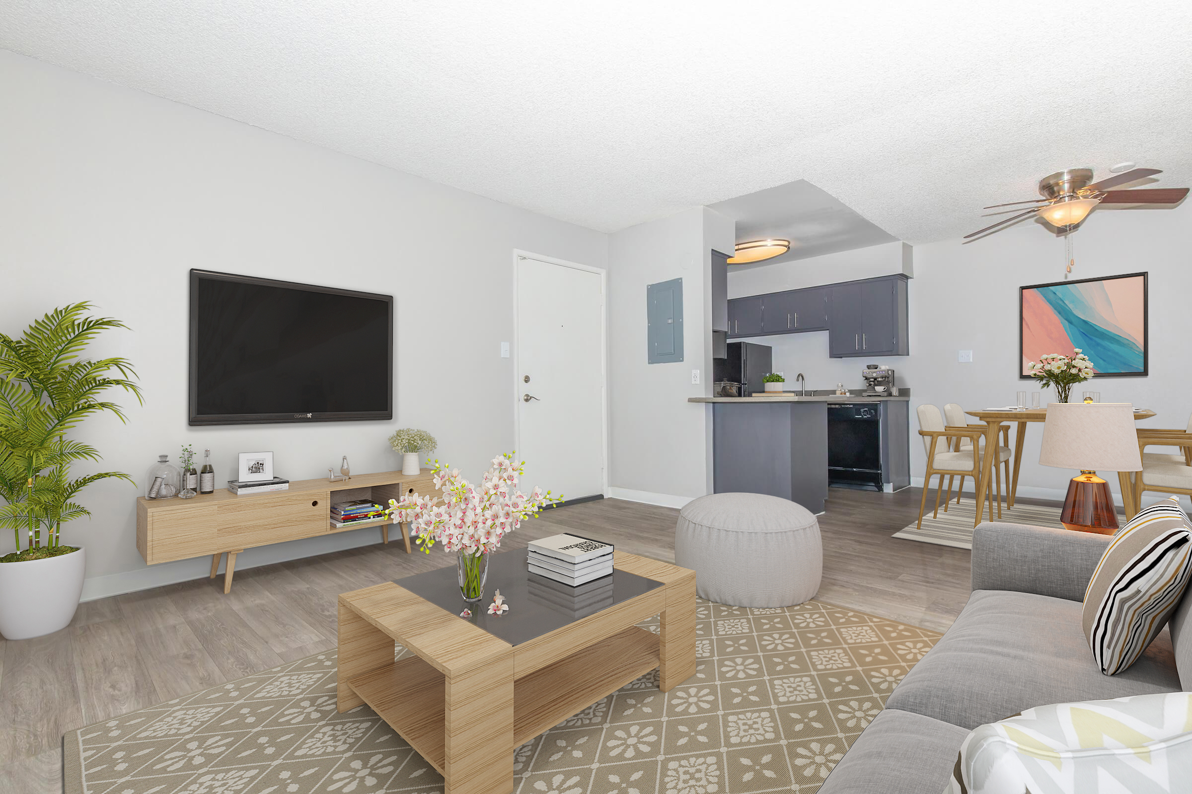 A modern living room featuring a gray sofa with patterned cushions, a wooden coffee table with decorative items, a large flat-screen TV mounted on the wall, and a potted plant. In the background, there is a dining area with a wooden table and four chairs, alongside a kitchen space. Bright natural light enhances the cozy atmosphere.