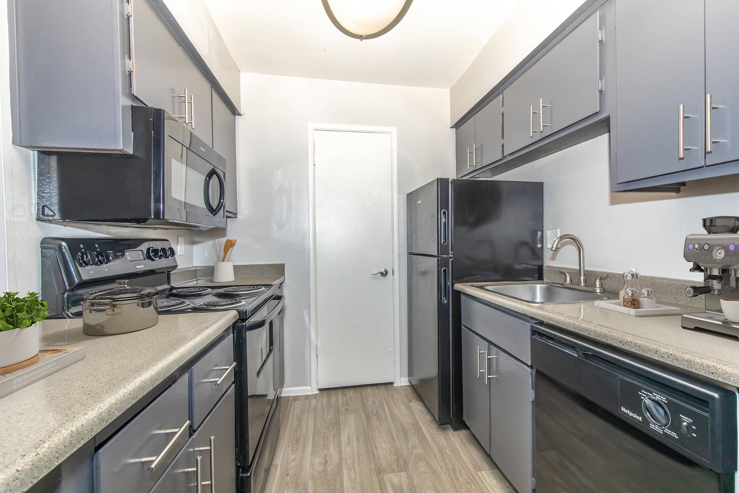 A modern kitchen featuring gray cabinets and appliances. It includes a stainless steel sink, a stovetop, an oven, and a refrigerator. The countertops are light-colored, with minimal decor, and a door leading to another area is visible in the background.