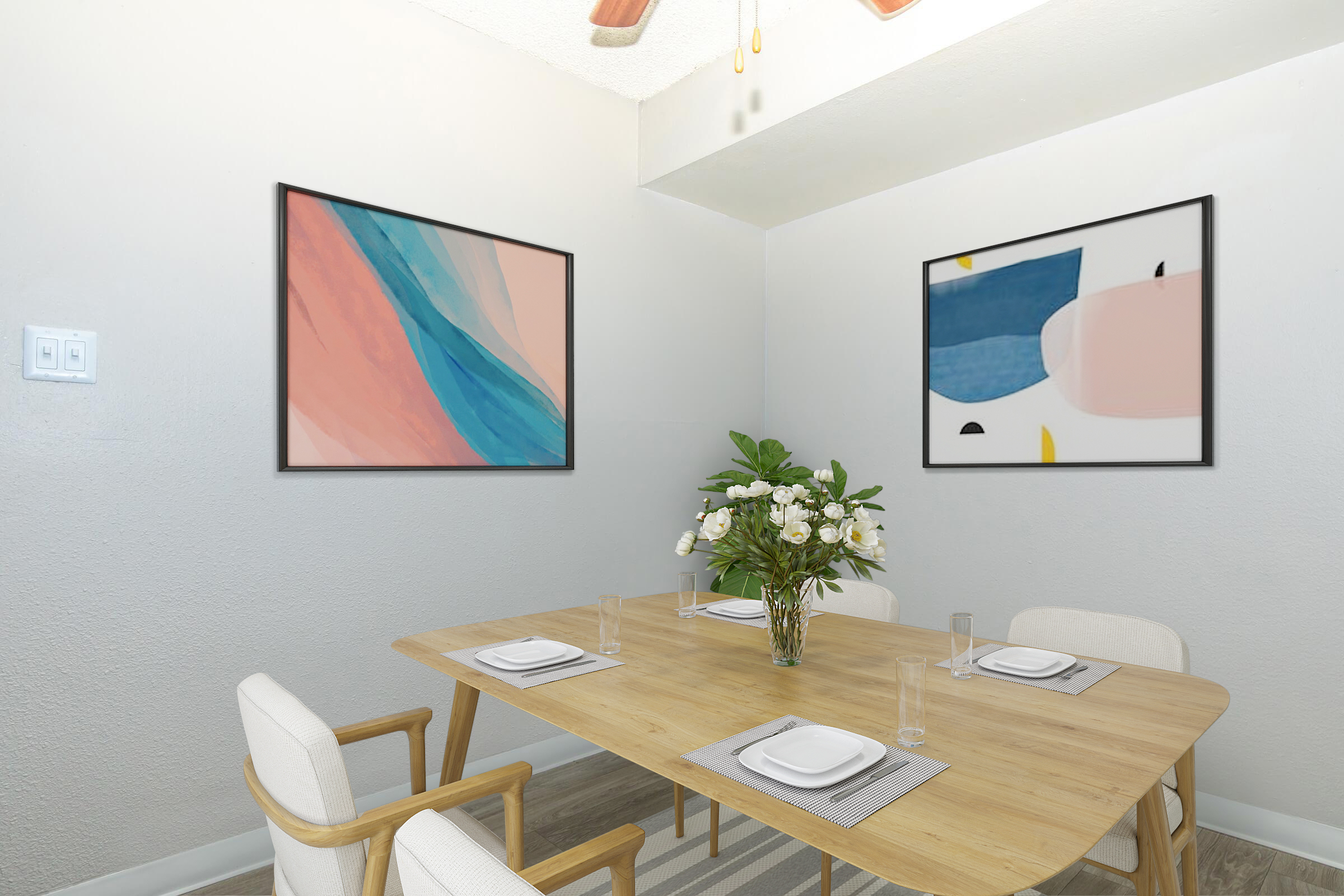 A modern dining area featuring a wooden table set for four. The table is adorned with white plates and glasses. Two abstract paintings hang on the light gray walls, with soft colors. A vase of fresh white flowers sits in the center of the table, complemented by stylish light-colored chairs.