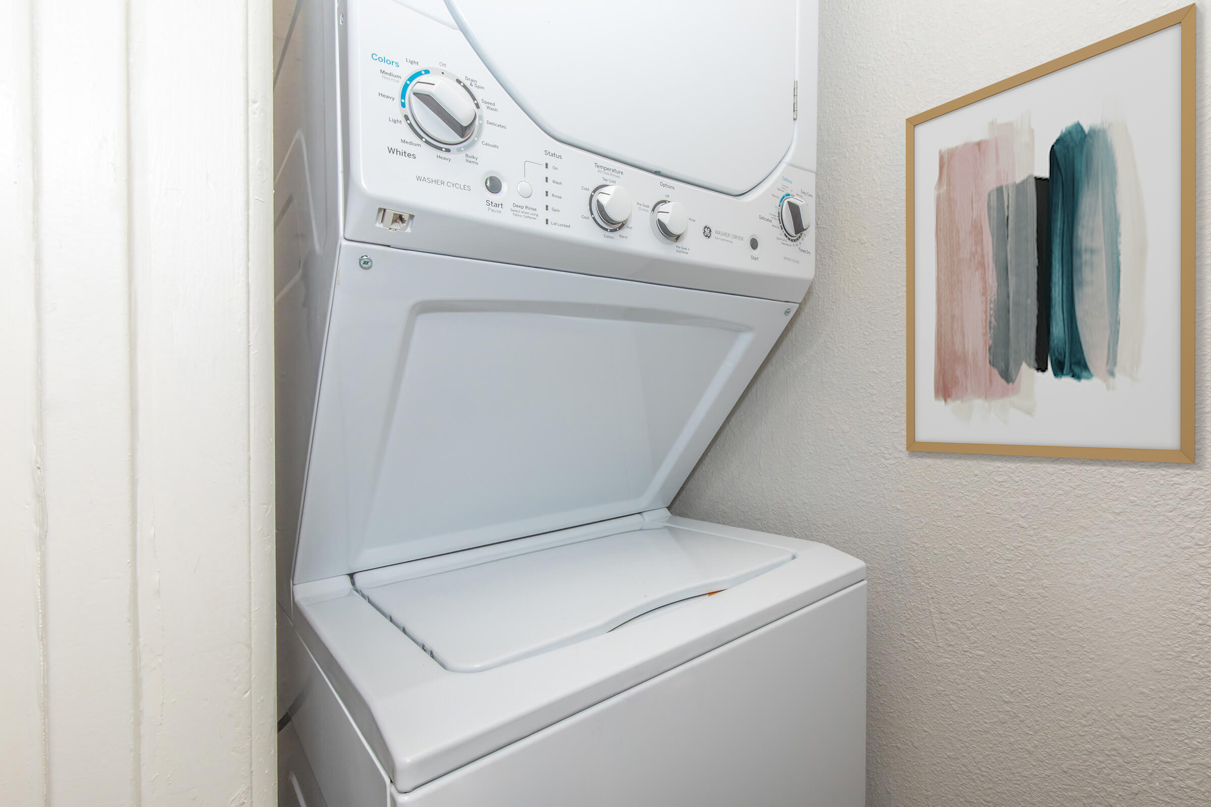 A stacked washer and dryer unit located in a small utility space, with a framed abstract artwork featuring soft colors on the wall next to it. The area has plain white walls and appears clean and organized.