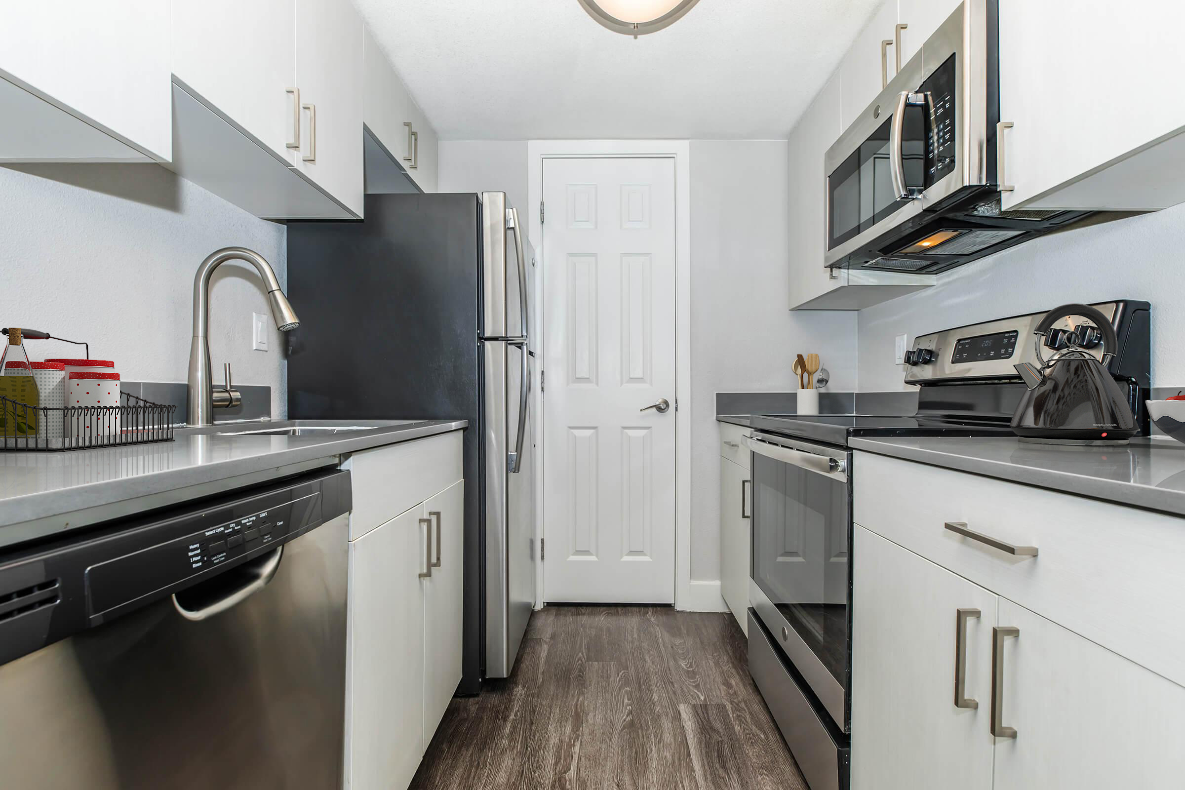 Modern kitchen featuring stainless steel appliances including a refrigerator, microwave, and oven. The cabinetry is light-colored with sleek handles, and the countertop is gray. A dishwasher is located next to the sink, with a door leading to another room at the end of the kitchen. The flooring is a brown laminate.