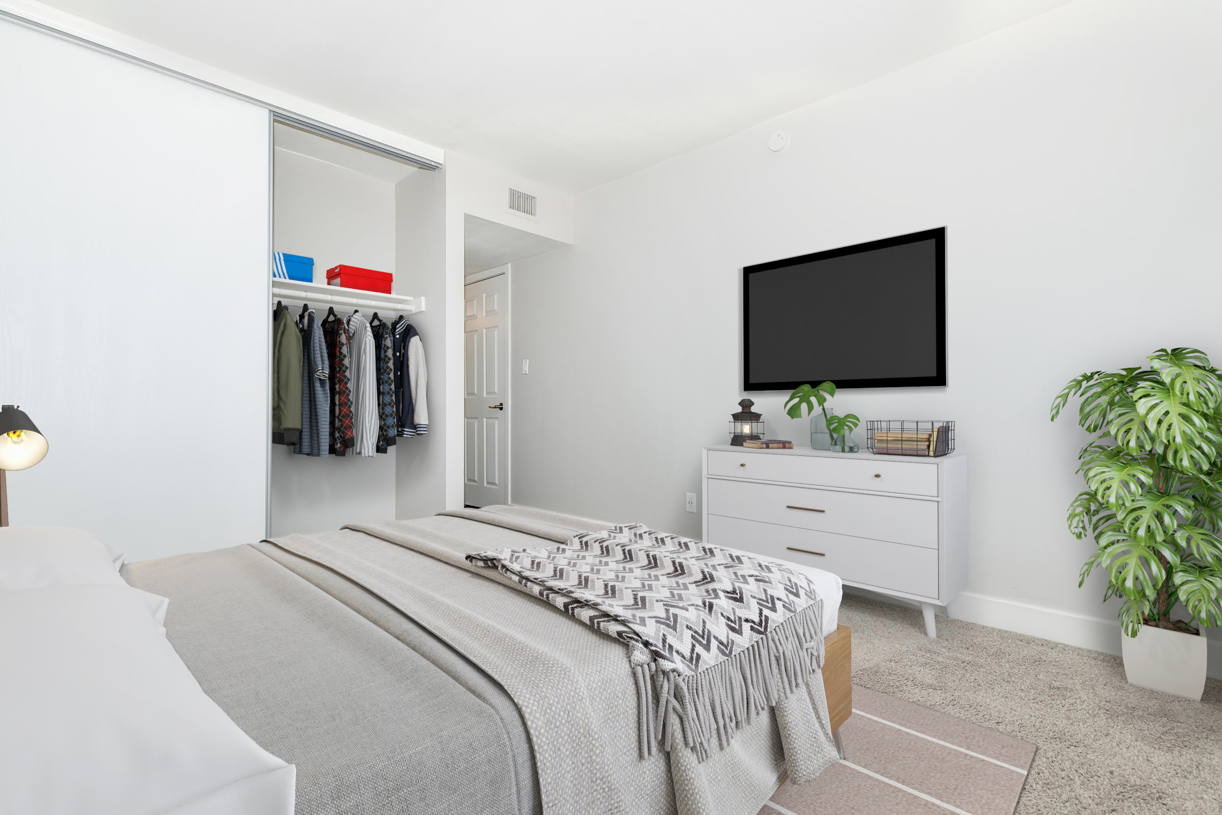 A modern bedroom featuring a neatly made bed with gray and white bedding, a closet with hanging clothes, a dresser with a decorative plant and books, a large wall-mounted TV, and a cozy area rug. The walls are painted white, creating a bright and airy atmosphere.