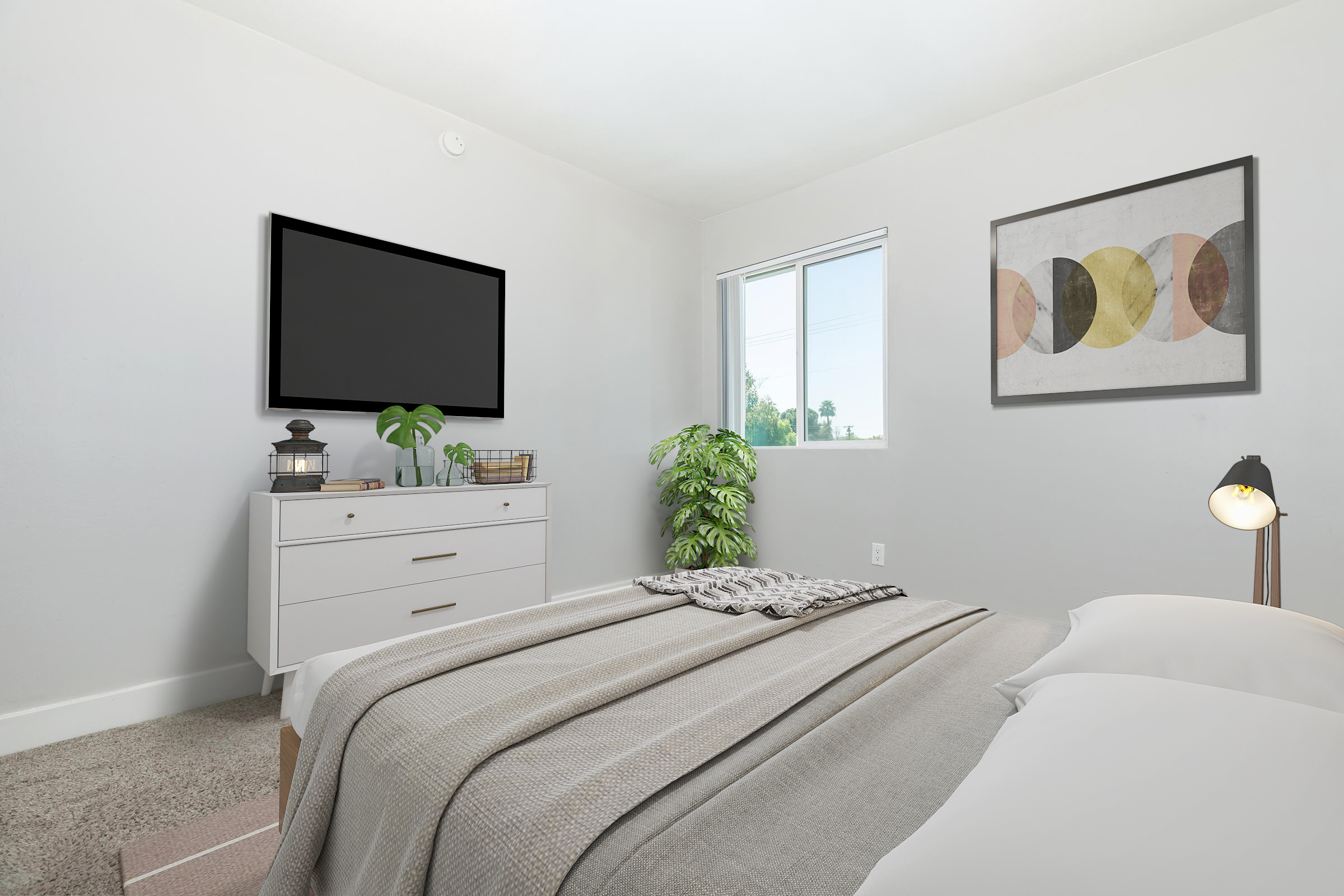 Brightly lit bedroom featuring a neatly made bed with white linens and a light gray throw. A dresser with decorative items is against one wall, while a large wall-mounted TV and a framed artwork adorn another. A small indoor plant adds a touch of greenery, complementing the minimalistic decor.