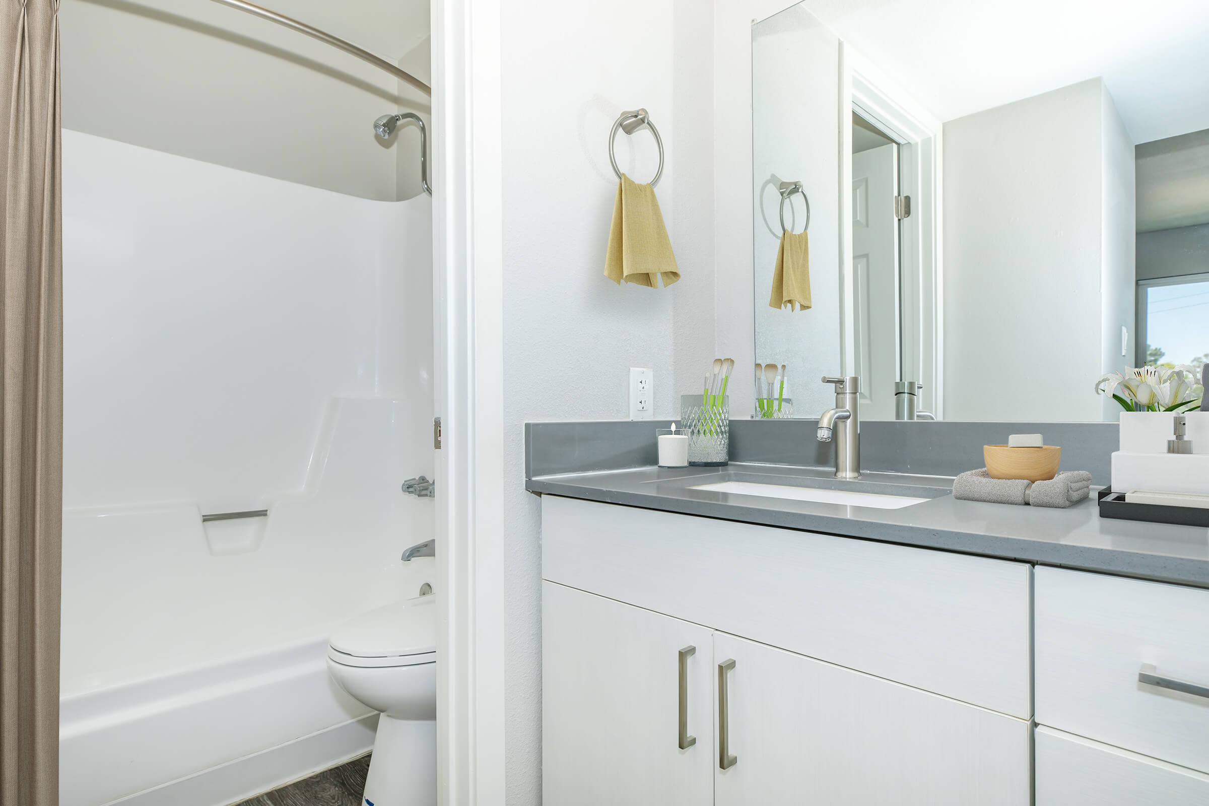 Modern bathroom featuring a shower with a curtain, a toilet, and a stylish sink with a gray countertop. The space is well-lit, with a mirror above the sink and decorative items like towels and a small vase of flowers on the counter. Clean and contemporary design elements create a fresh atmosphere.