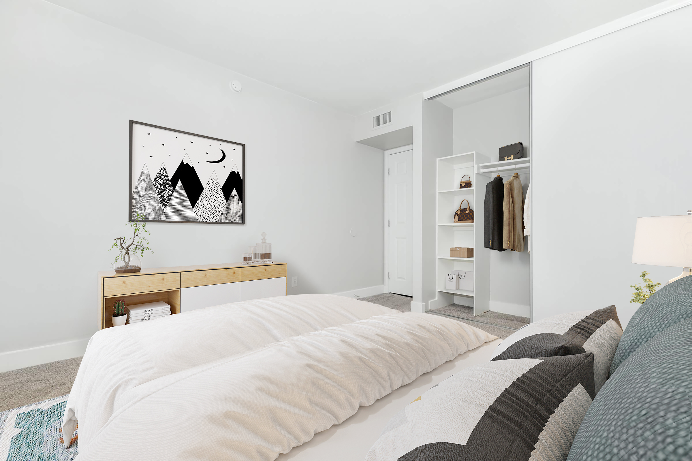 A cozy bedroom featuring a large bed with white bedding and decorative pillows. There's a framed black and white mountain artwork on the wall. To the right, a closet with hanging clothes is visible, and a wooden dresser sits at the foot of the bed. Natural light brightens the space, enhancing the minimalist decor.