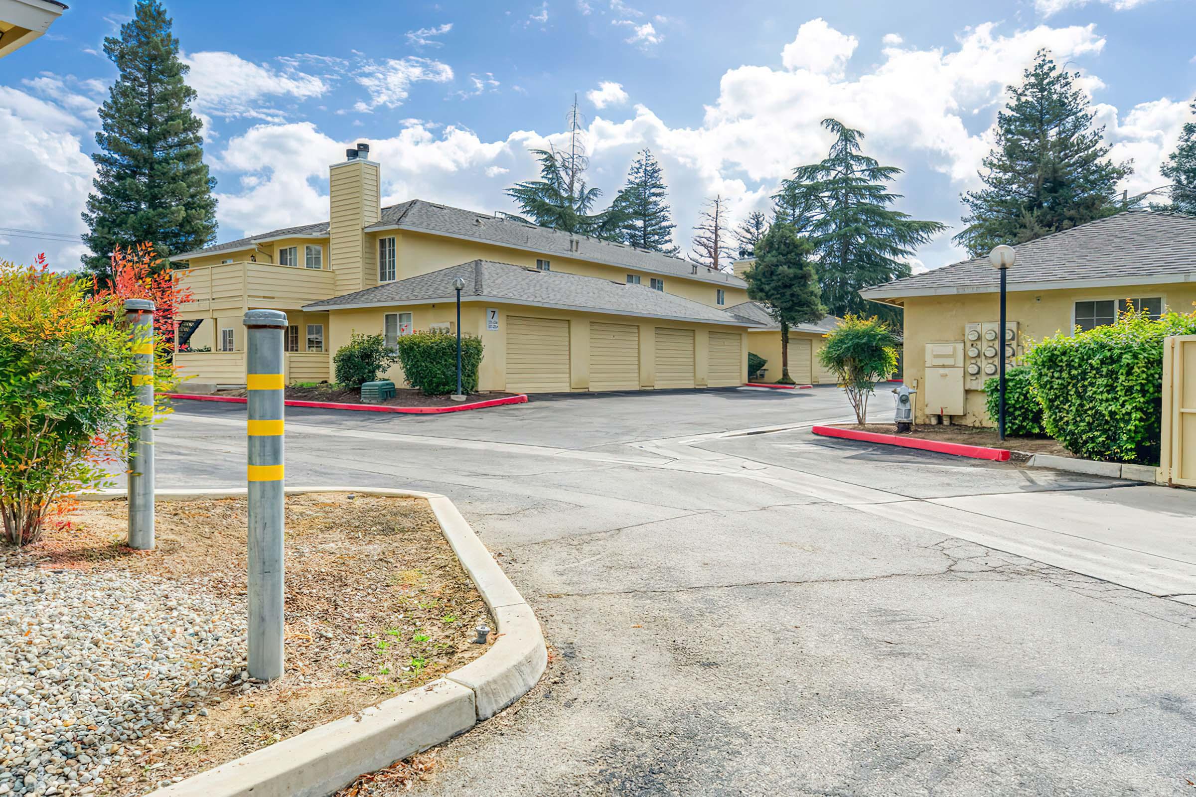 A residential complex featuring two-story buildings with beige siding, surrounded by green trees and shrubs. A paved driveway leads to garages, with a red-bordered parking area. The sky is partly cloudy, creating a bright atmosphere.
