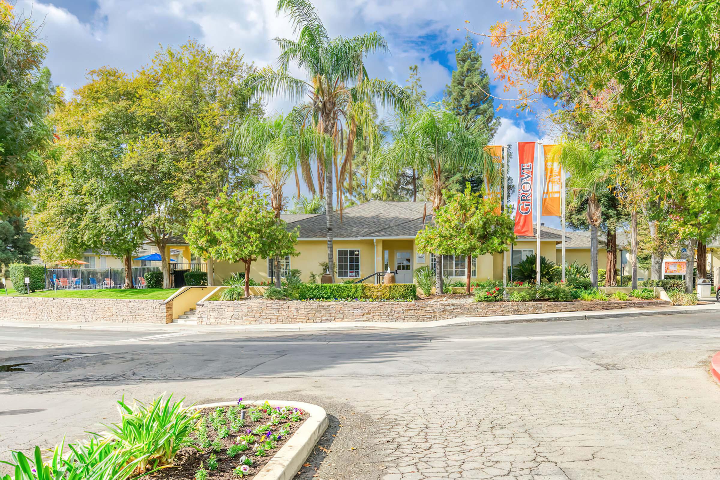 A welcoming entrance to a property featuring a beige building surrounded by lush greenery, palm trees, and colorful banners. The landscape includes flower beds and a winding road leading up to the entrance, creating a warm and inviting atmosphere.