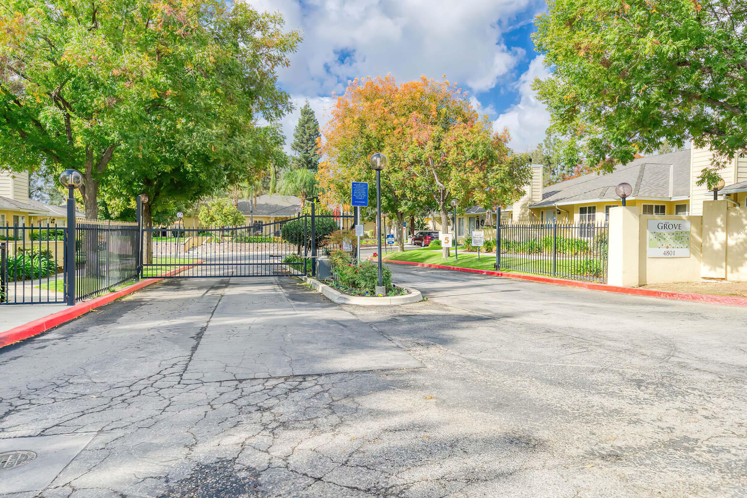 A gated entrance to a residential community, featuring a tall gate, trees with autumn foliage, and a sign labeled "Grove." The road is cracked and leads into the complex, with cloudy blue skies overhead. Nearby, there are well-maintained sidewalks and landscaping.