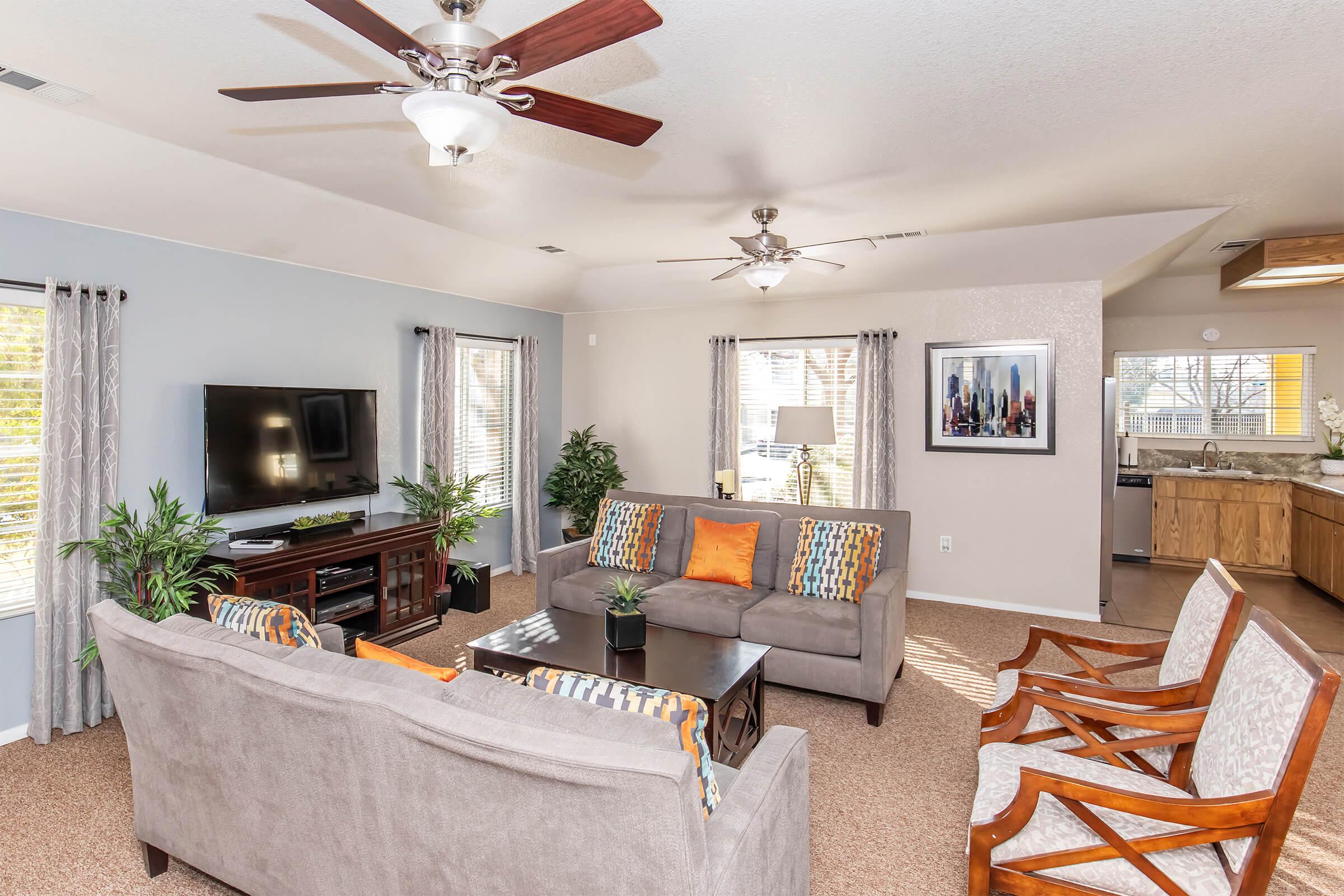 Bright and inviting living room featuring a gray sofa with orange and patterned cushions, two wooden chairs, a TV on a stand, and large windows with light curtains. Potted plants add a touch of greenery, and the space opens to a kitchen area in the background.